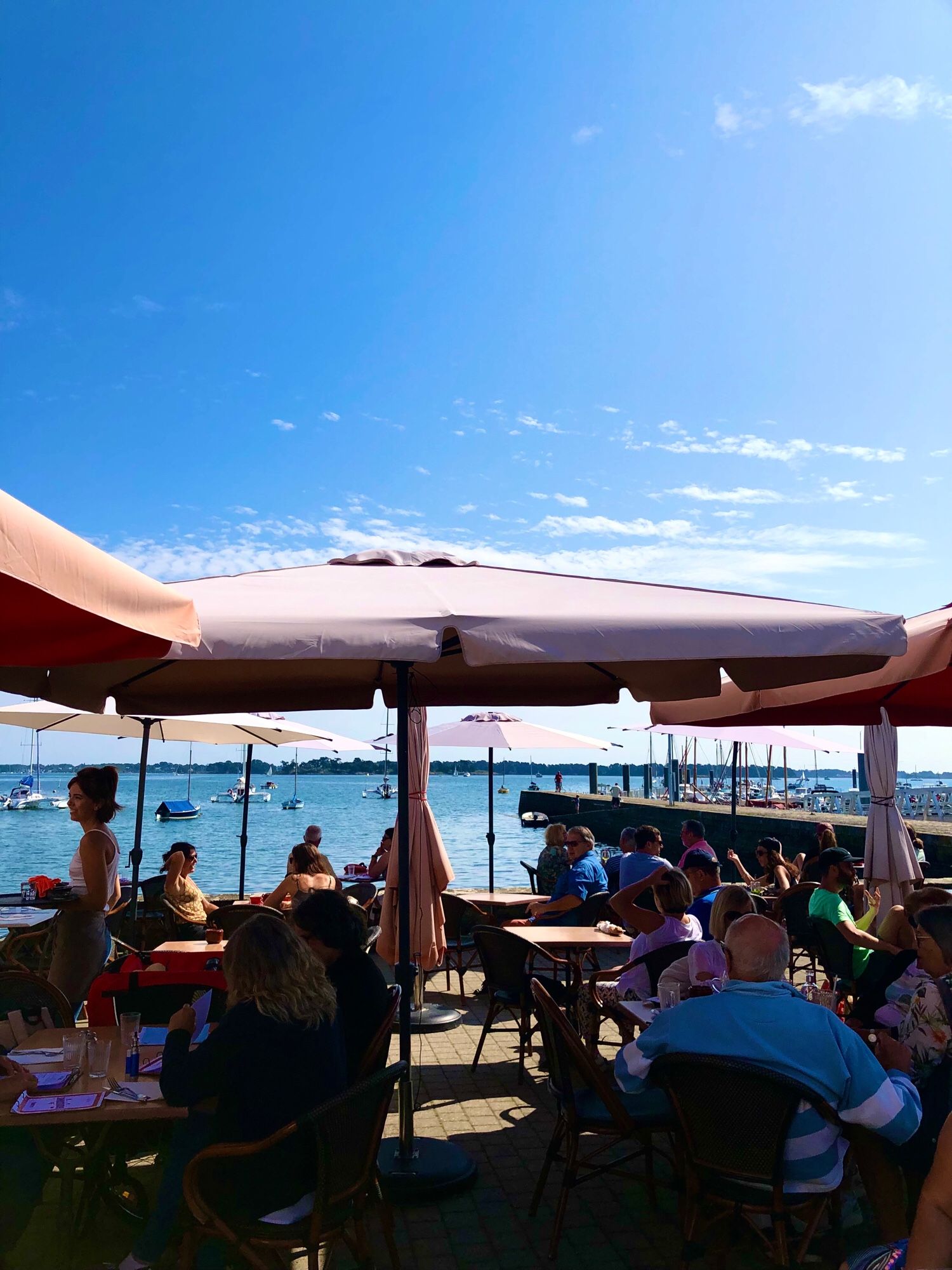 Café en terrasse avec des parasols oranges blanchis par le soleil, beaucoup de monde, vue sur mer bleue et ciel un peu nuageux