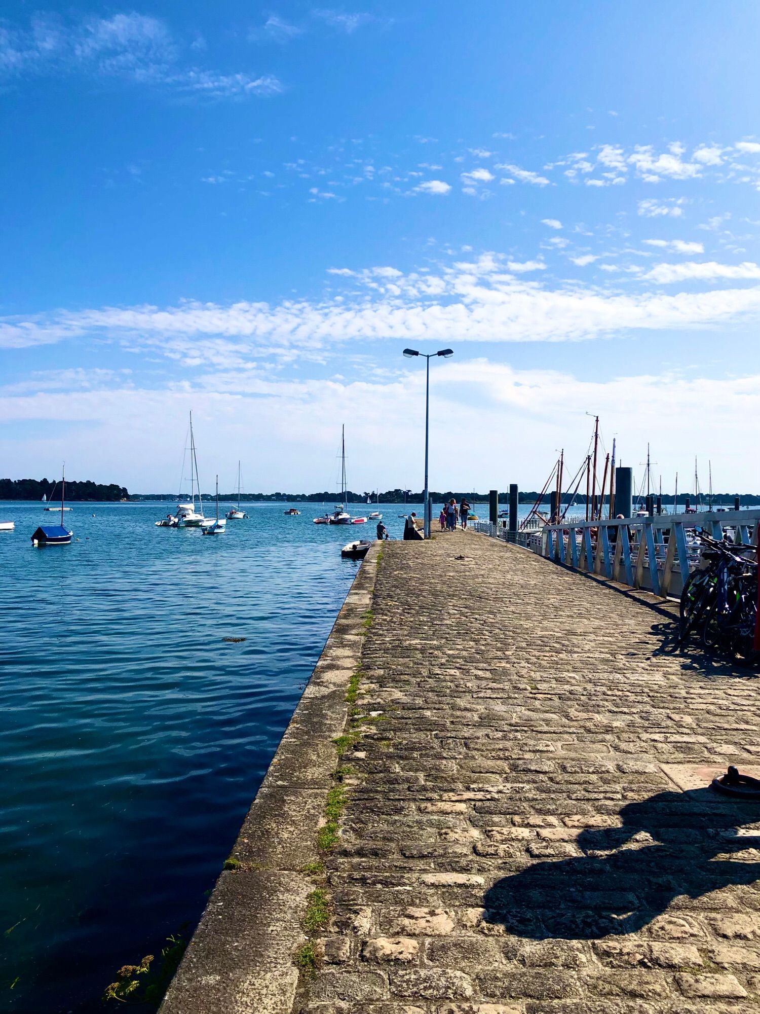 Petit port avec digue mer bleue et ombres de 2 personnes en bas à droite, qq bateaux dans le fond