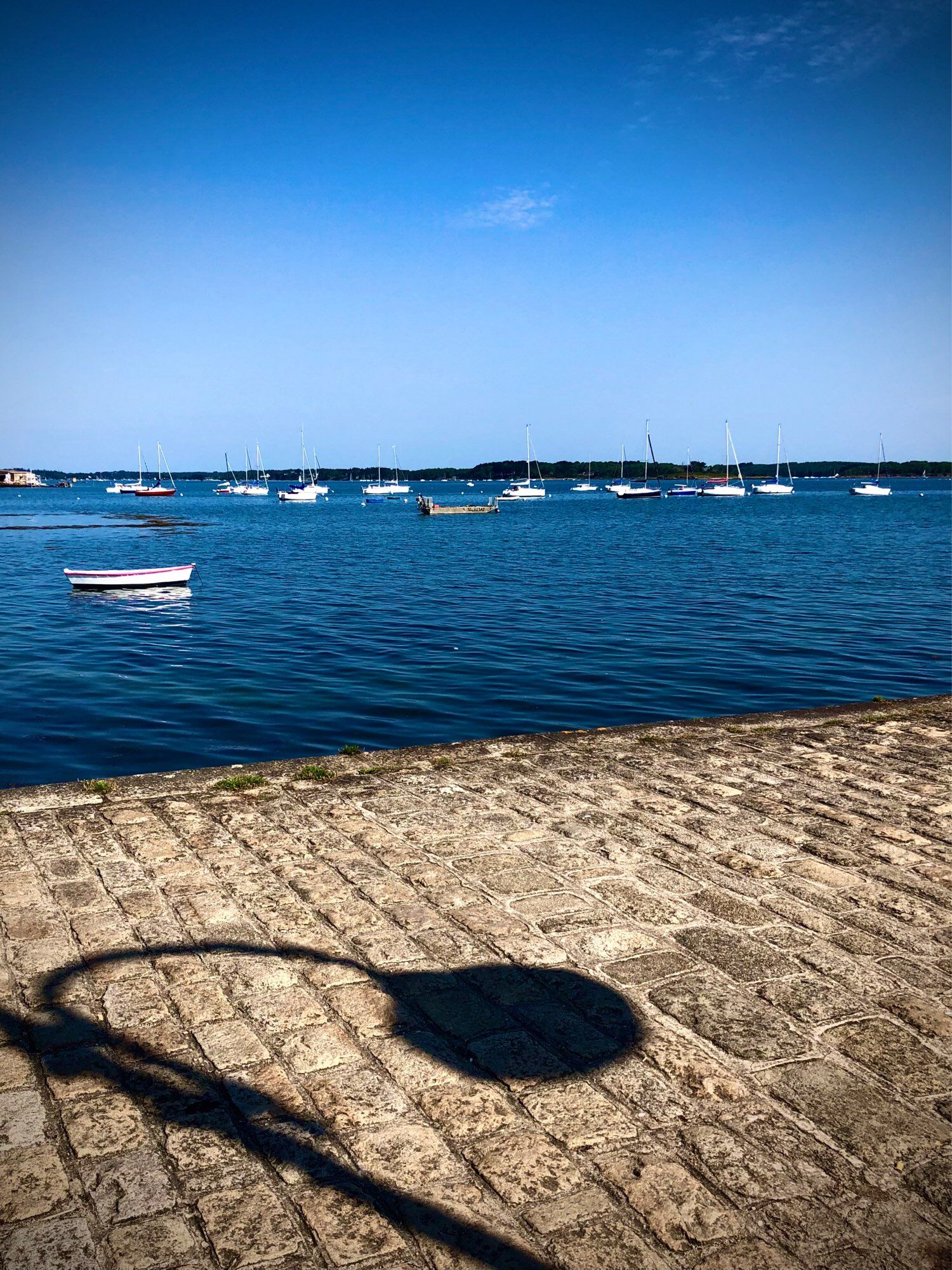 Ombre d’un réverbère sur une digue, barque sur l’eau bleue et plein d’autres dans le loin, ciel bleu