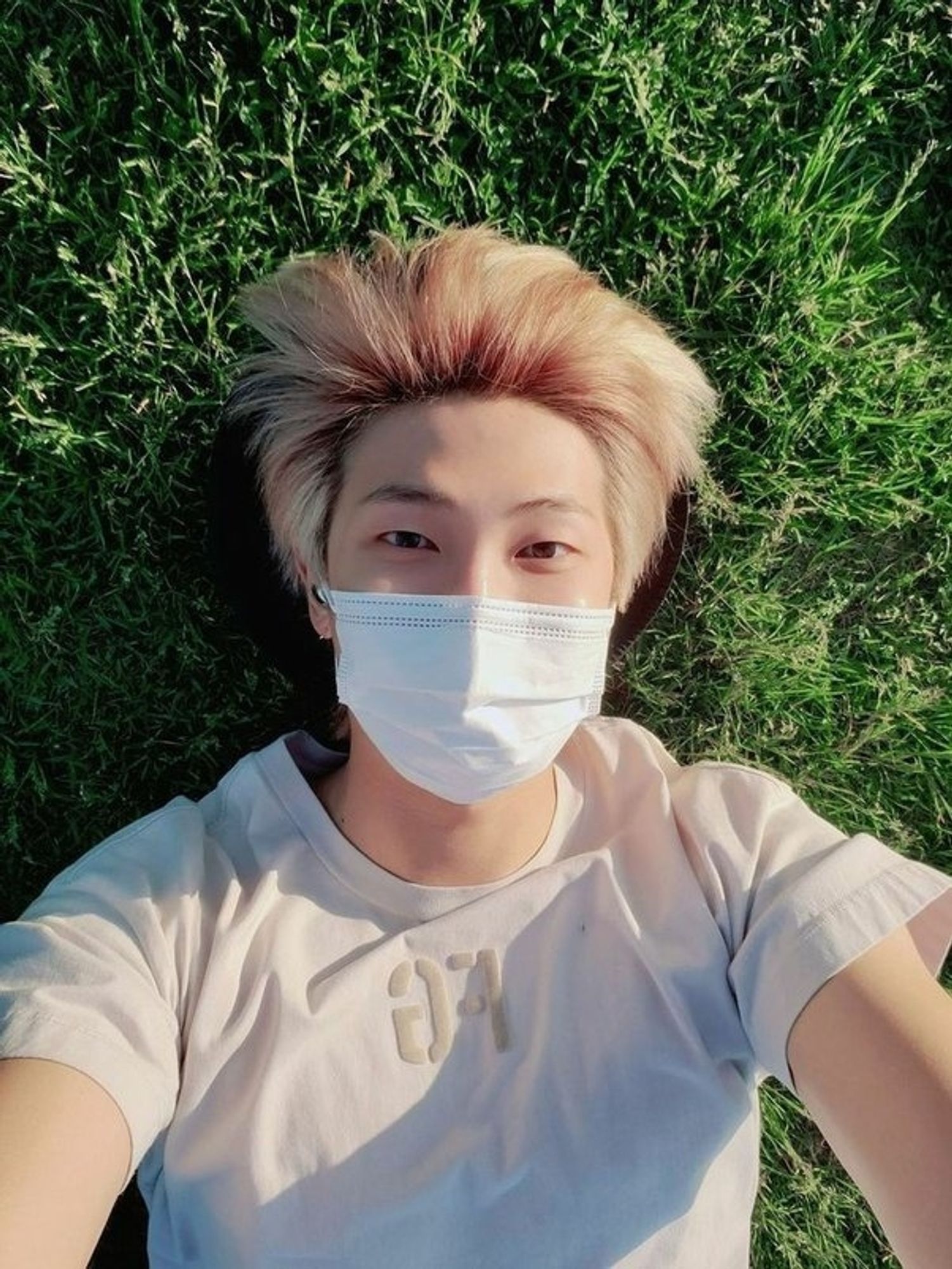 Namjoon laying on the grass, blond and with a cream colored shirt.