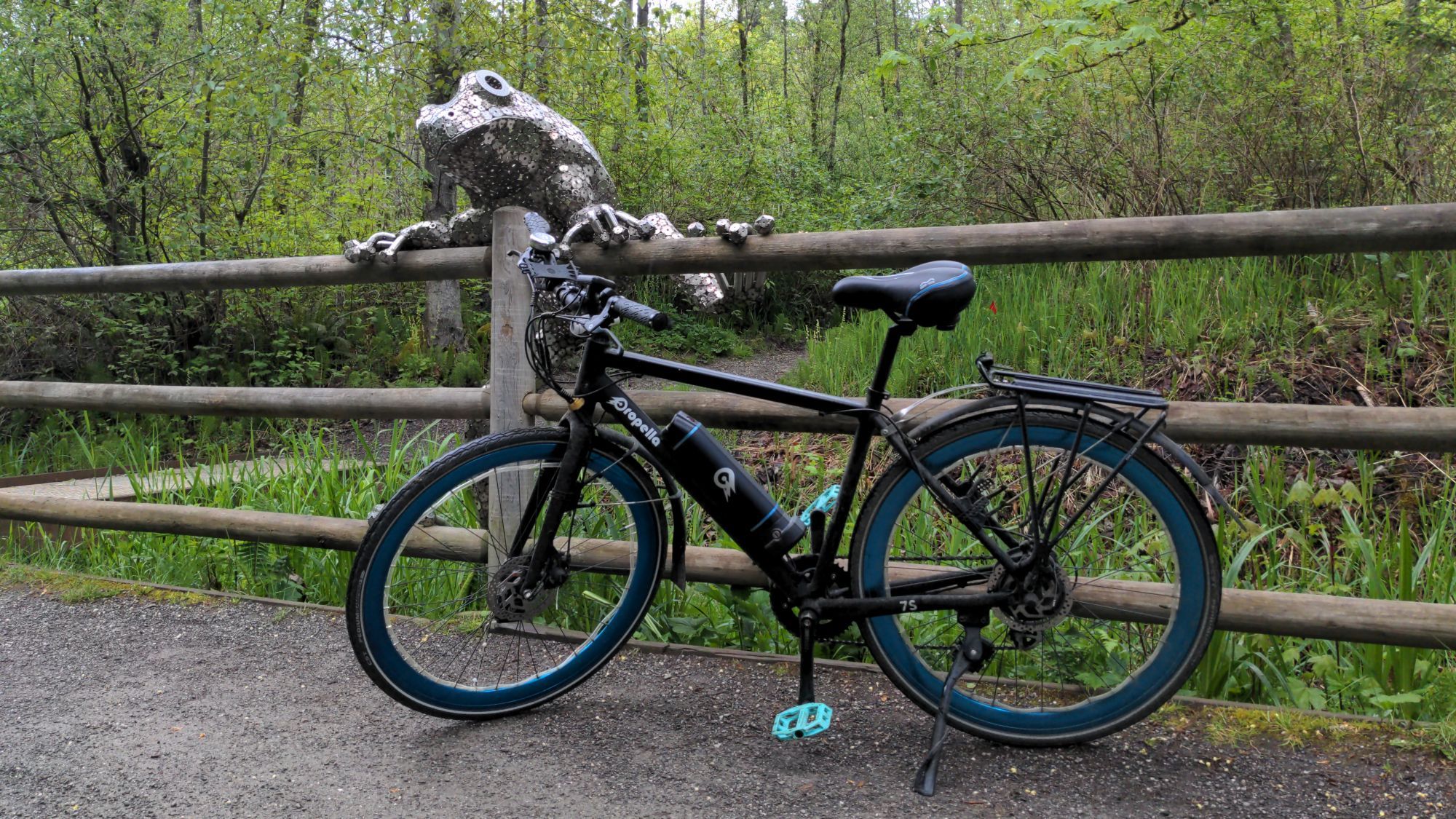 A Propella 7S Class 1 ebikr with electric Blue rims and bright Teal pedals sits on a gravel trail in front of a wooden fence, and the foliage and lush greenery behind it. On the fence is a metal sculpture of a frog, climbing over the top rail an wooden post. The sculture is made of tiny interconnected metal circular discs, resembling that or construction washers, welded and joined together - with gaps between them, rendering the interior hollow. The Sculture is named Frog Xing by Will Schlough, placed in 2022 on the Cross Kirkland Corridor.