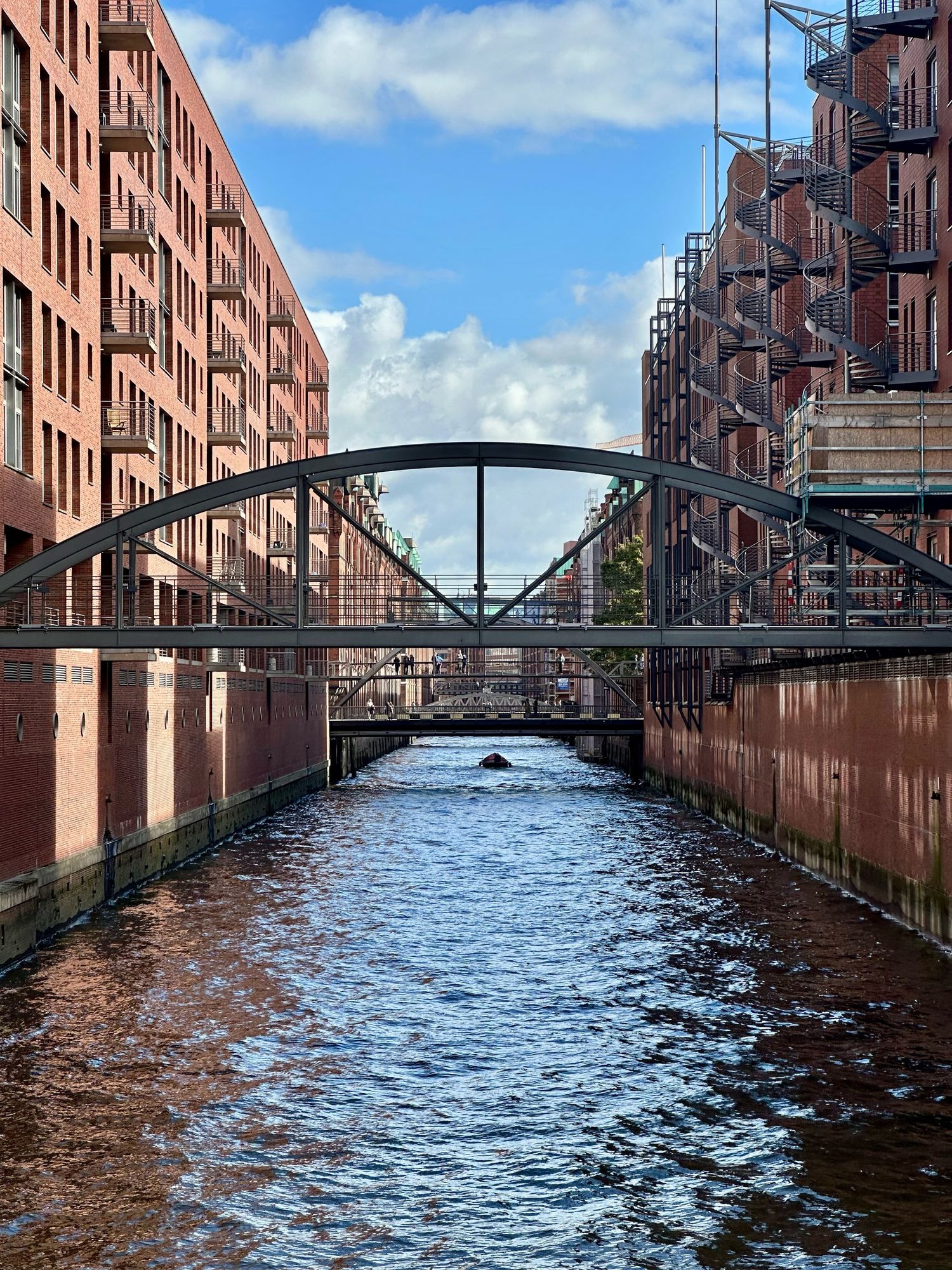 Blick über einen Kanal der Speicherstadt. Links und rechts Backsteinhäuser, mittig Brücken. Weiter weg im Kanal ein Boot.