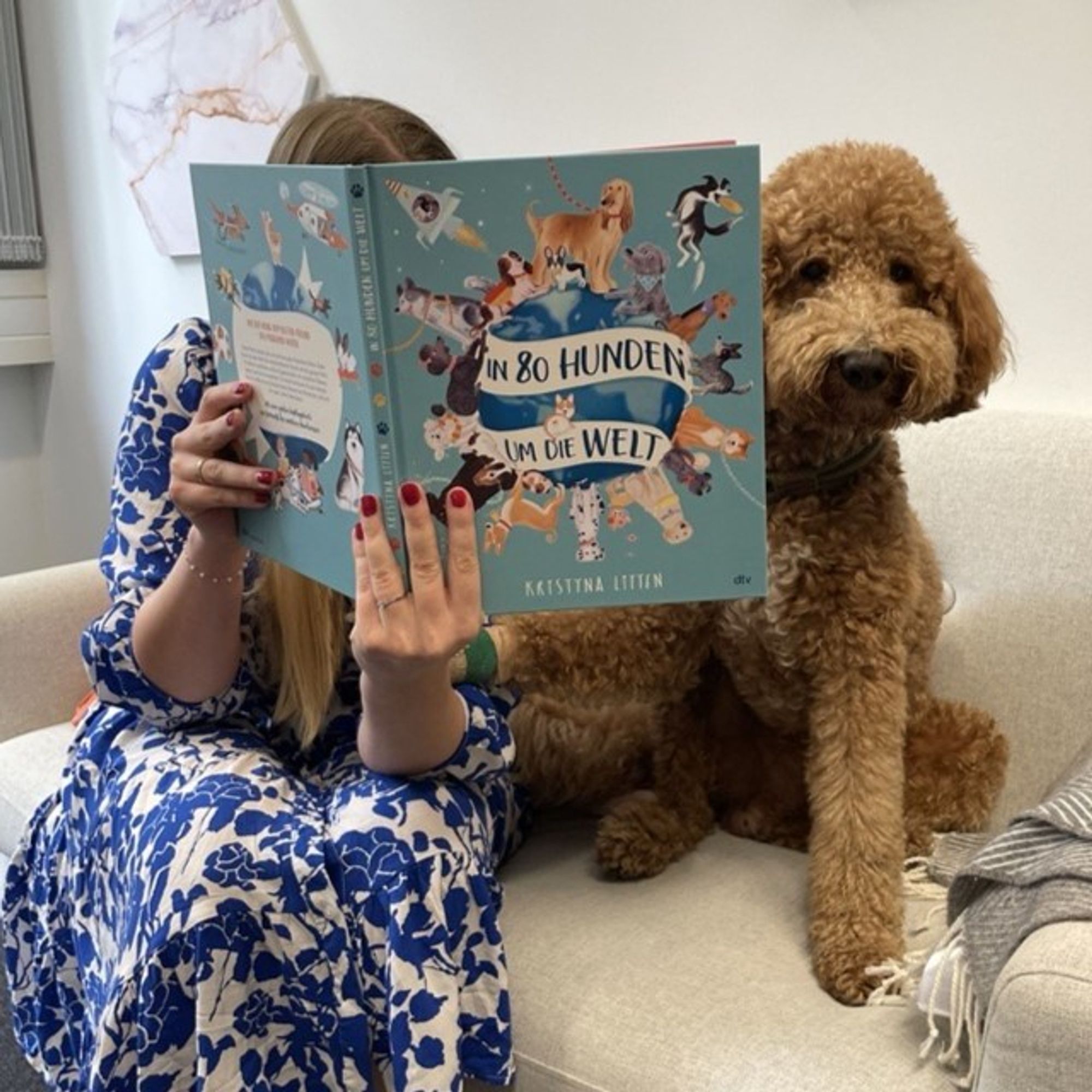 Eine Frau sitzt zusammen mit dem Goldendoodle Hans auf dem Sofa und liest mit ihm das Buch "In 80 Hunden um die Welt". Der Hund schaut hinter dem Buch hervor in die Kamera