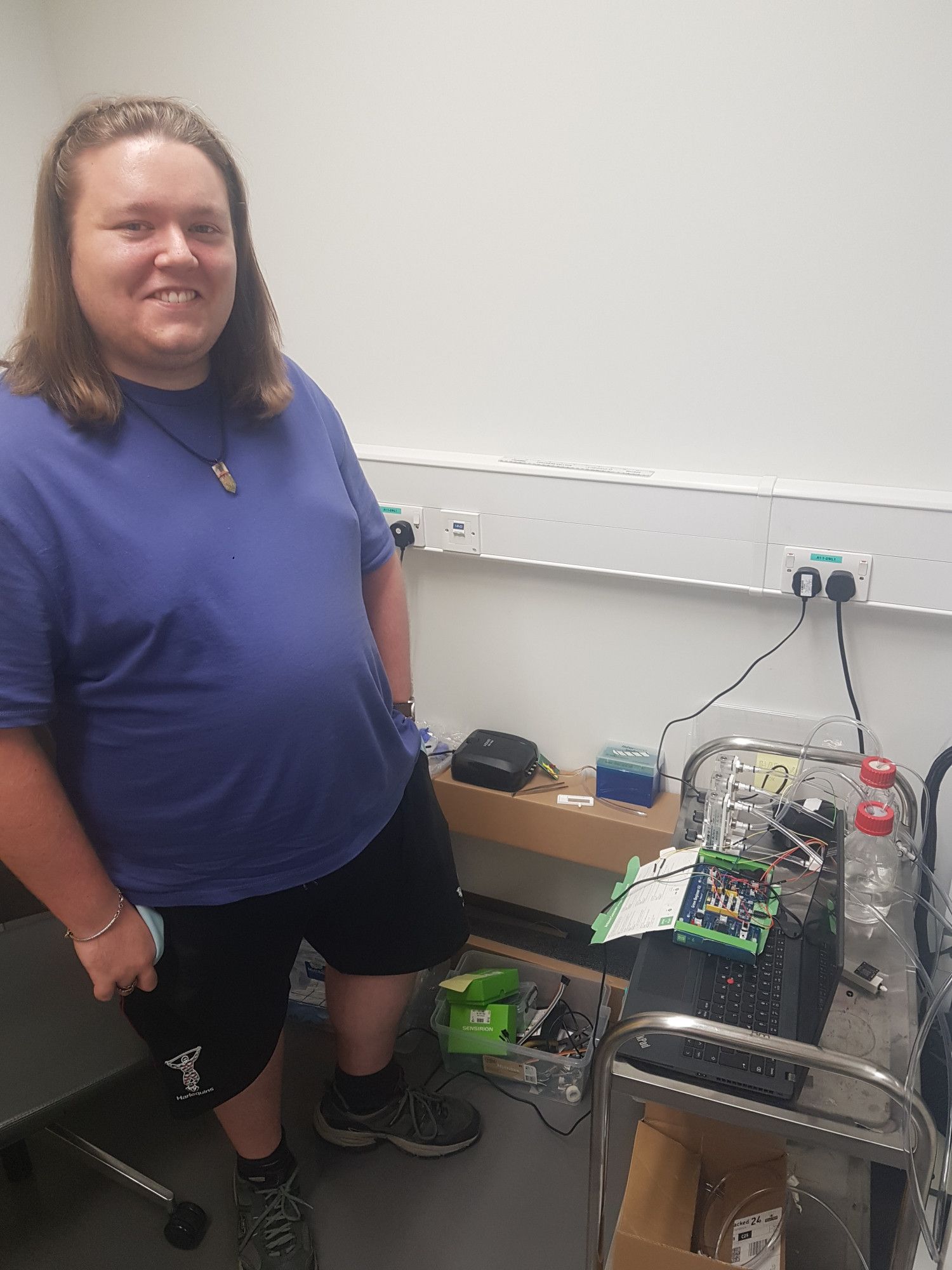 Long haired man in blue T shirt and black shorts standing next to a collection of electronics, tubing and bottles in a lab.