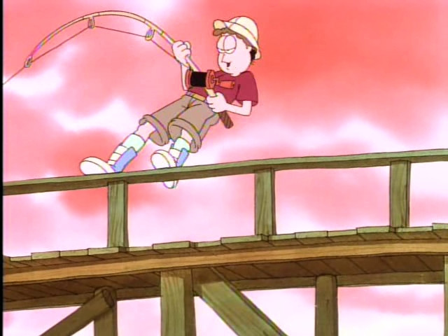 A screenshot from the Garfield & Friends TV show. Jon Arbuckle is standing on a pier fishing, in a red t-shirt and khaki shorts/capris. He is wearing light blue high top shoes and knee length white socks. Jon appears determined, reeling in a big fish and bracing himself against the handrail on the pier with his feet and standing upright against the tension. The fishing pole is bent nearly in half.