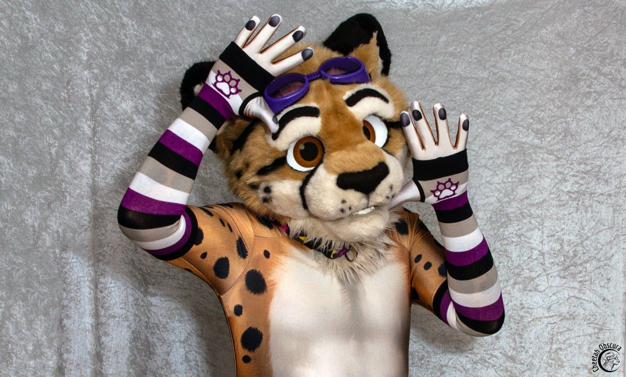 Photo of Obscura in fursuit partial, spandex petsuit, and asexual color arm warmers