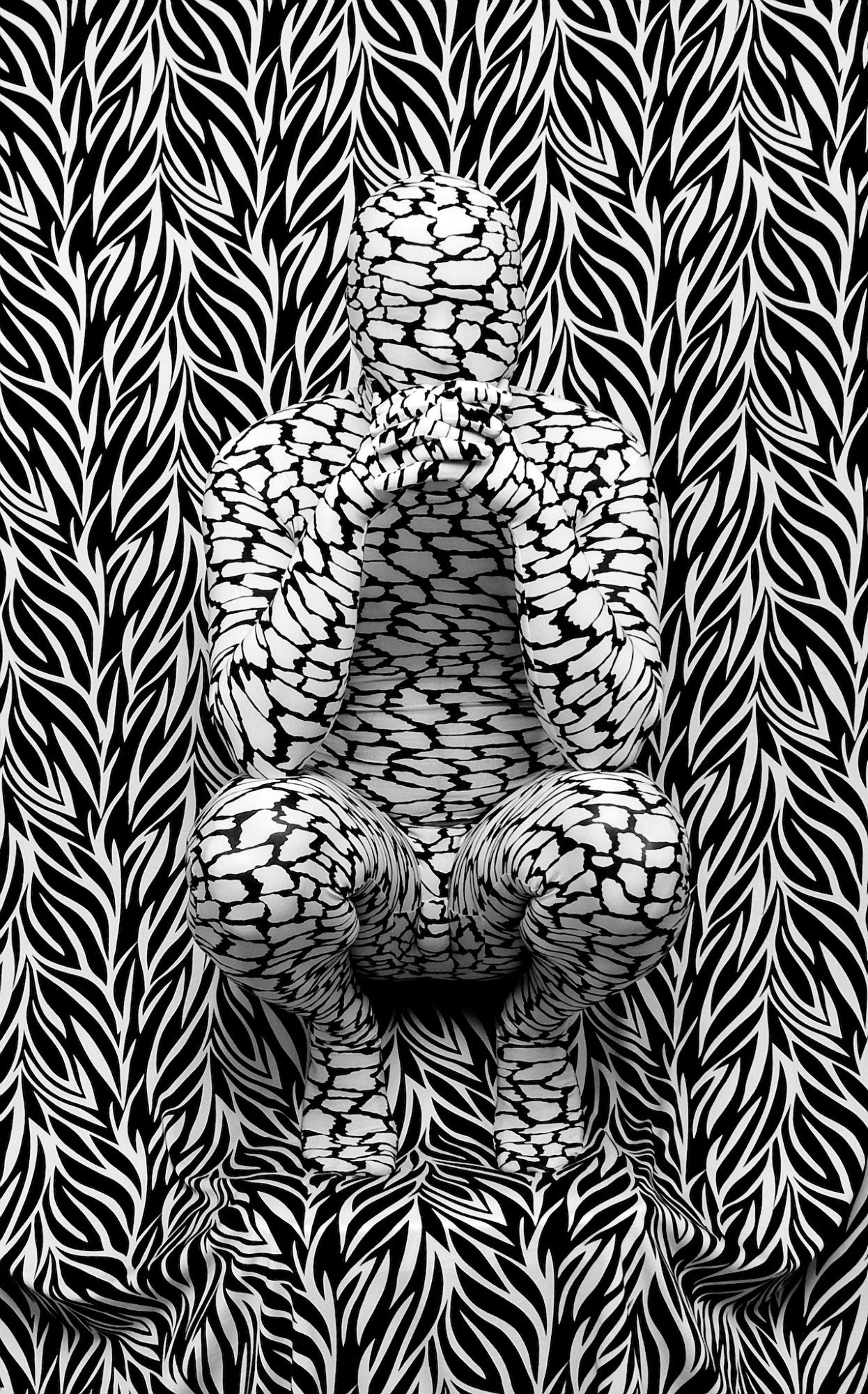 Black and white photo of a figure in a dry lake bed pattern zentai crouched in front of a zebra print background