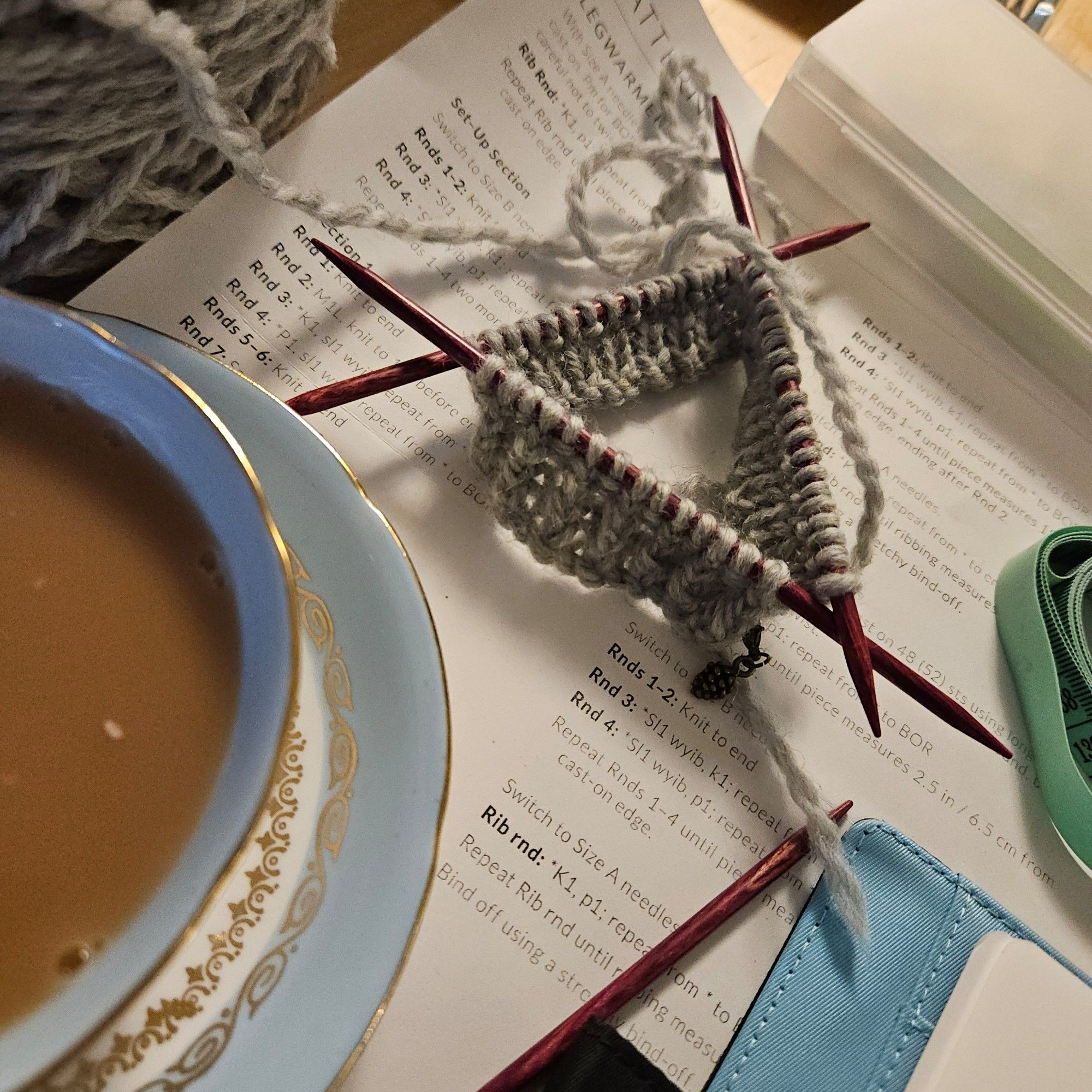 Grey yarn knit up in twisted rib stitch on three double pointed needles sits atop a pattern page. A cup of tea and general clutter frames the scene.