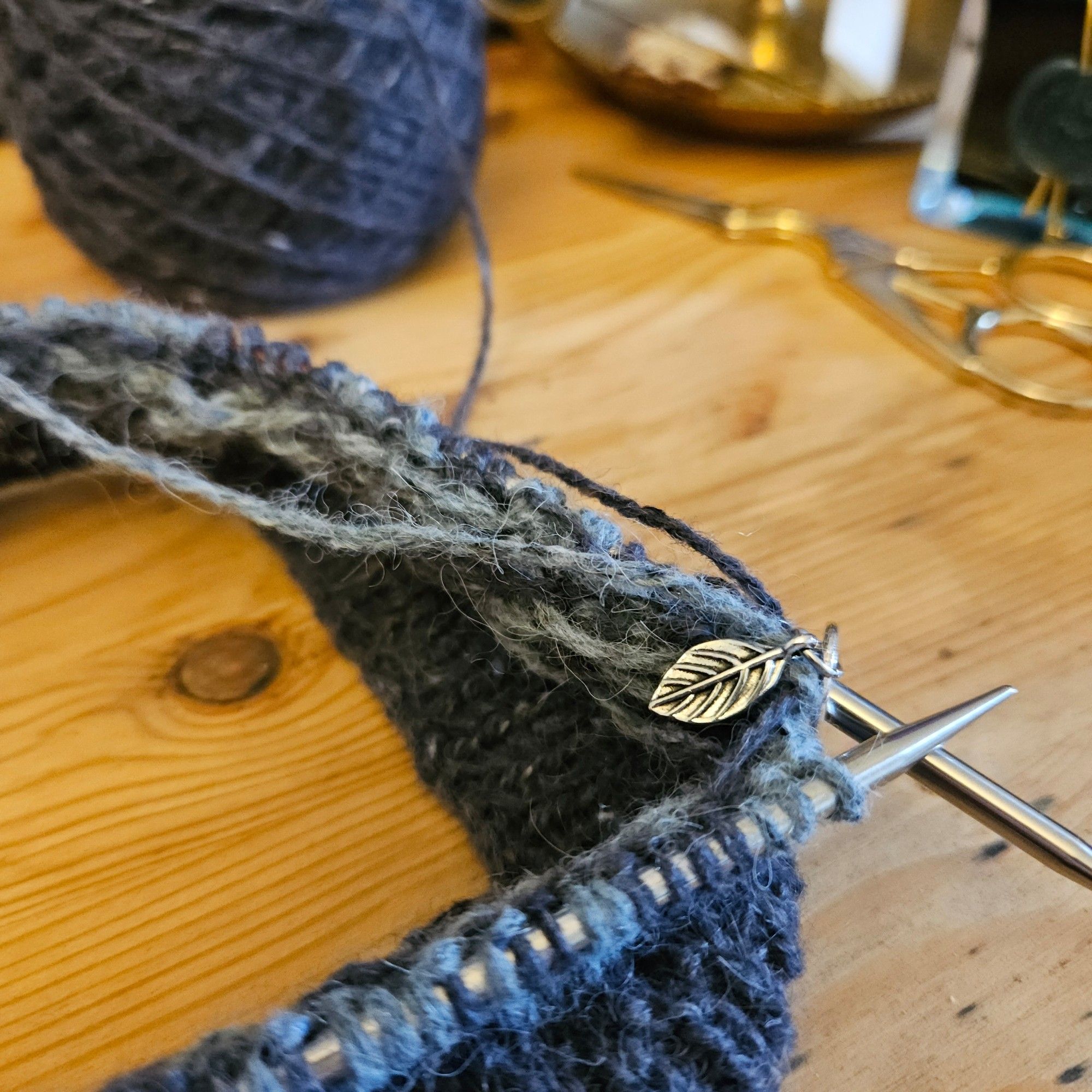 some knitting, black and blue tweed yarns, on circular needles, on a wooden desk. A small silver leaf stitch marker is in focus.