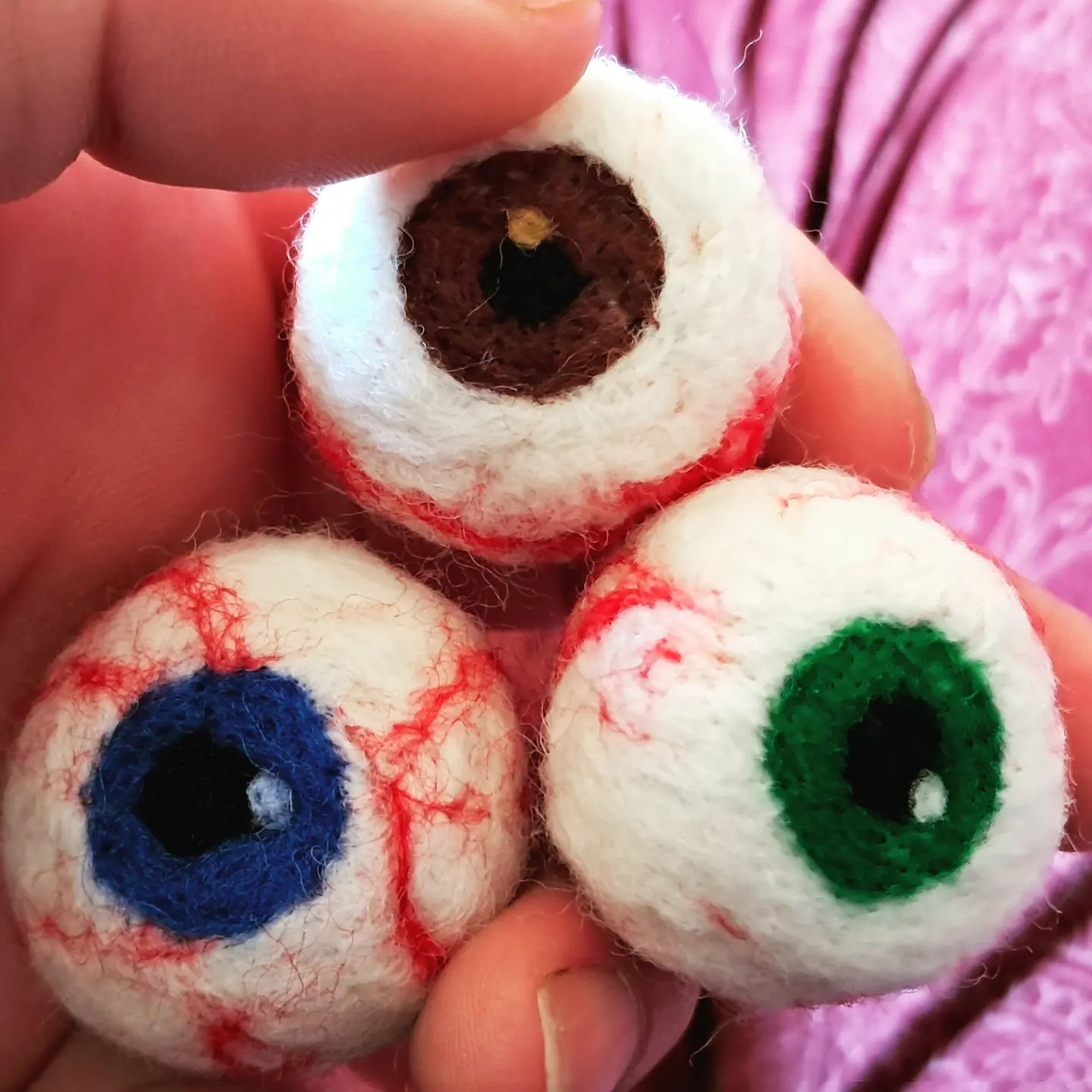 Three felted eyeballs held in a hand with brown, green, and blue irises