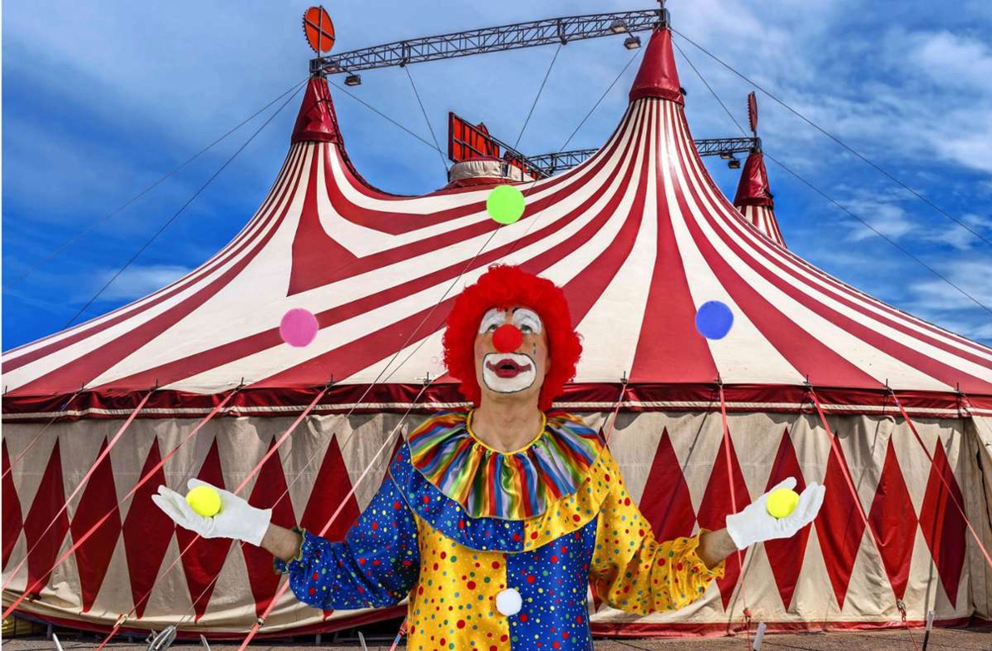 Das Bild zeigt einen Clown mit roter Perücke, roter Clownsnase und typischem weißem Gesicht mit buntem Make-up. Der Clown trägt ein buntes Kostüm mit Punkten und einem regenbogenfarbenen Kragen. Er jongliert mit mehreren bunten Bällen. Im Hintergrund ist ein großes rot-weiß gestreiftes Zirkuszelt zu sehen, das mit Seilen befestigt ist. Der Himmel ist klar und blau, was auf einen sonnigen Tag hindeutet.