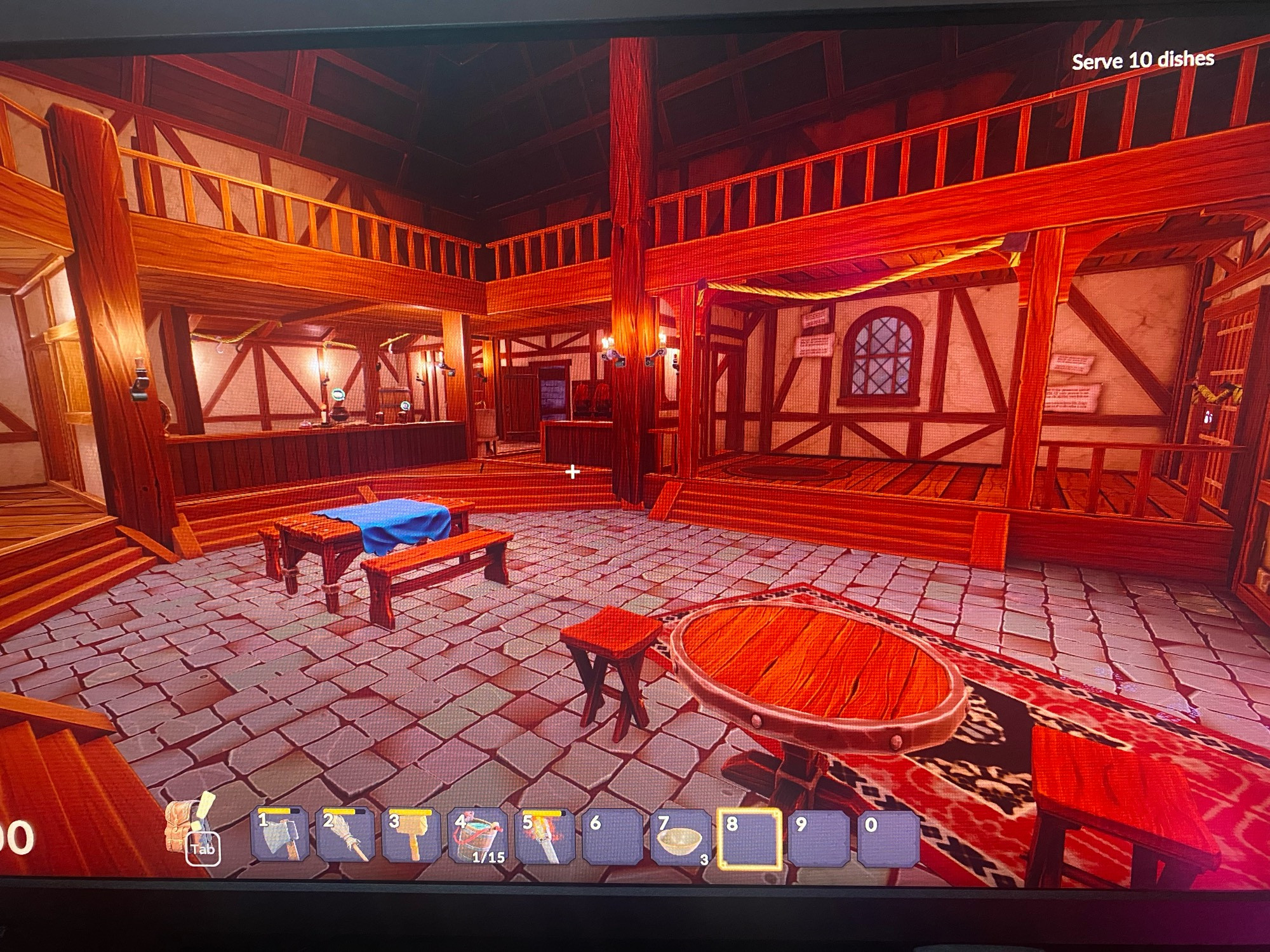 Image shows the interior of a medieval tavern in a cosy fantasy game, mounted candles light the room. The tables are on lower stone floor surrounded by raised wooden decks. The walls appear to have crossbeams. There is a second floor balcony above the wooden decks, overlooking the central dining area. The tavern has a warm and welcoming atmosphere despite being closed presently. 