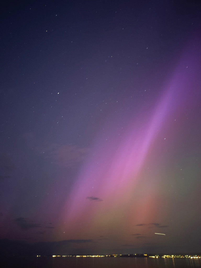 It's night and we're looking out over the sea to sparkly lights on the far shore.  Up in the sky are curtains of colour, pinks, greens and reds (the aurora) and some stary dots in the sky.