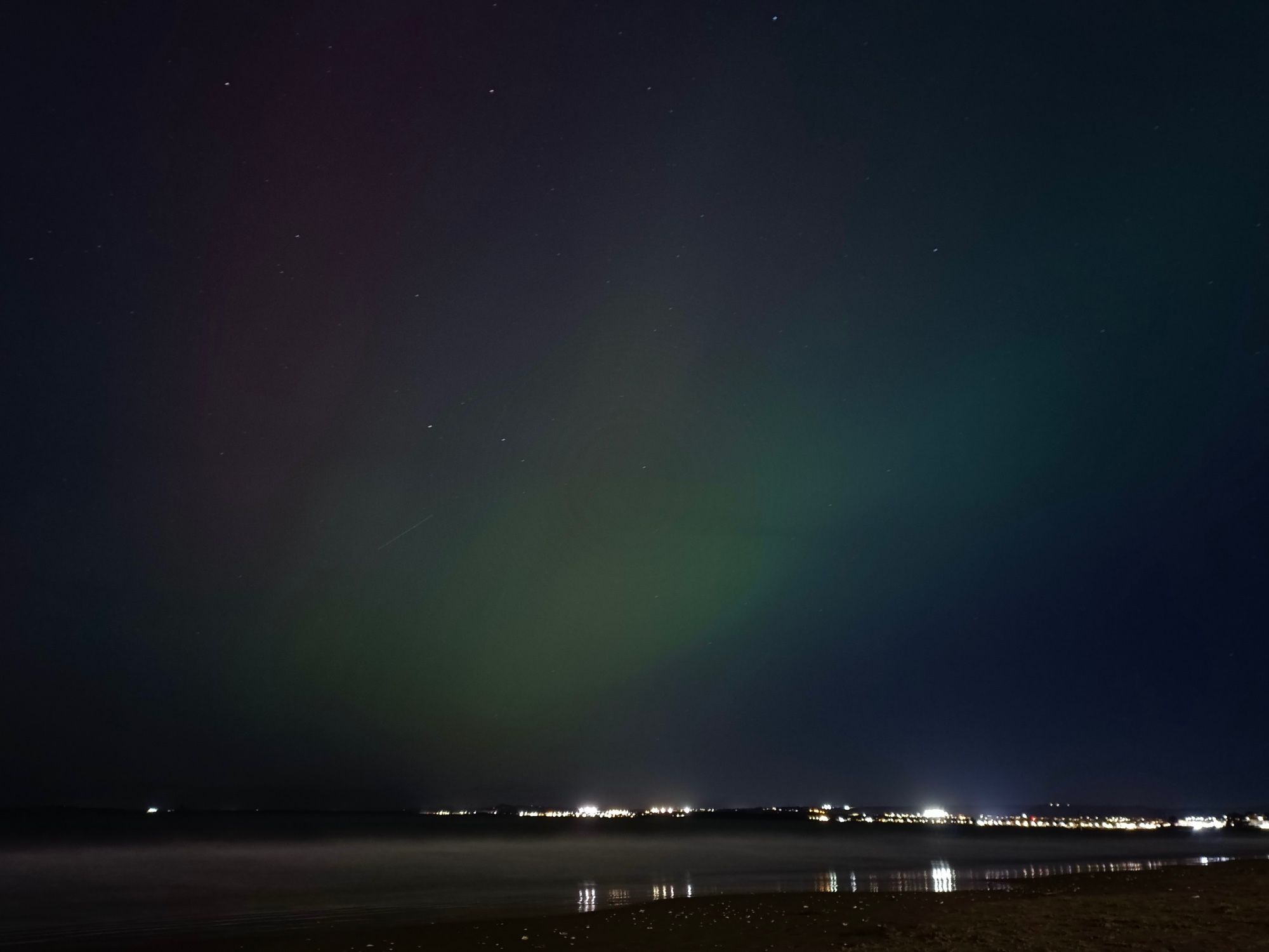 A beach at night time.  There are some cool rainbow colours in the sky, changing from red across to green.  Some stars dot the background