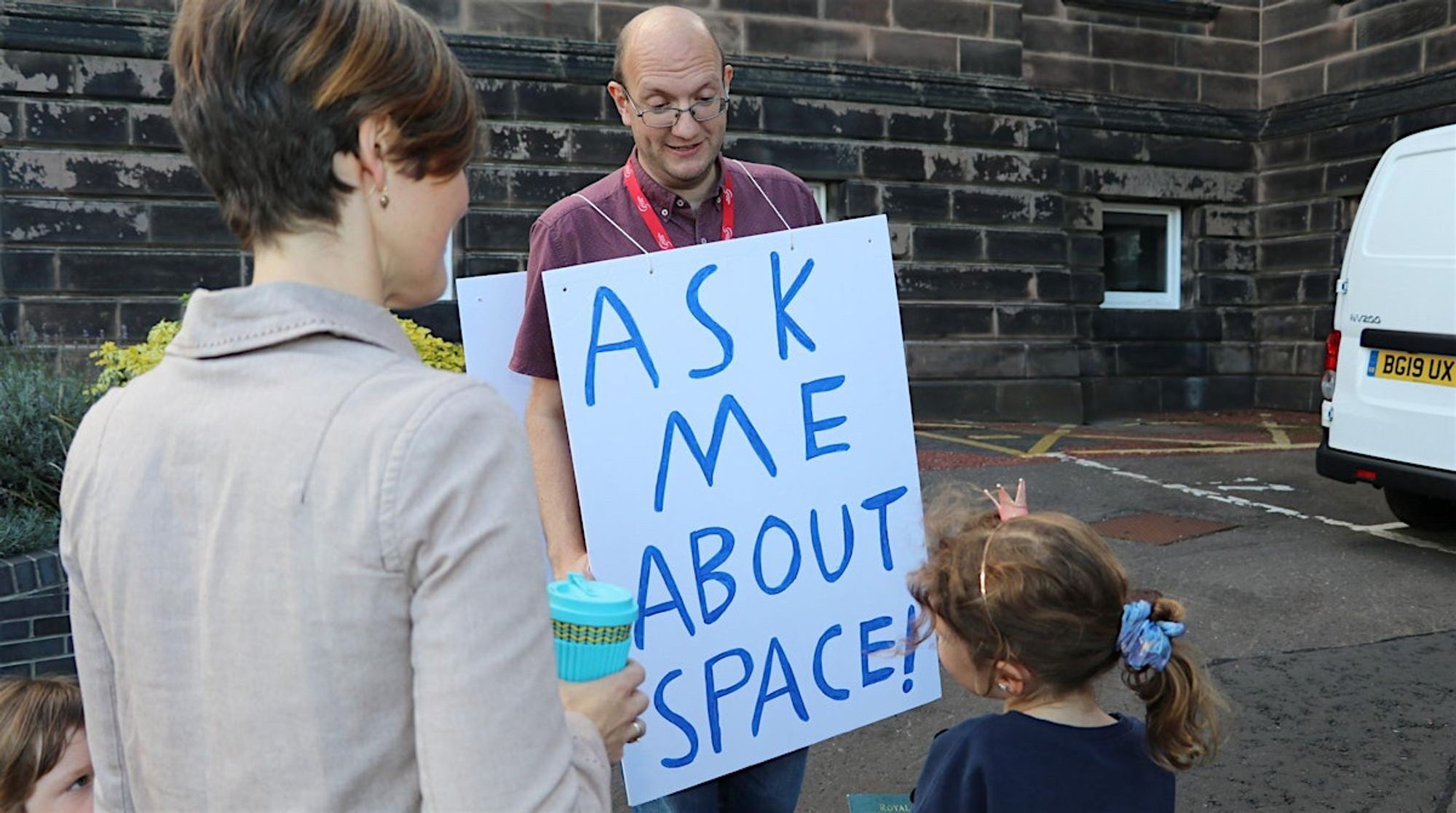 A small girl wearing an adorable tiny pink crown is looking up at a bald bespectacled man wearing a placard that says "Ask me about space!"  A Mum and little brother look on from the left.