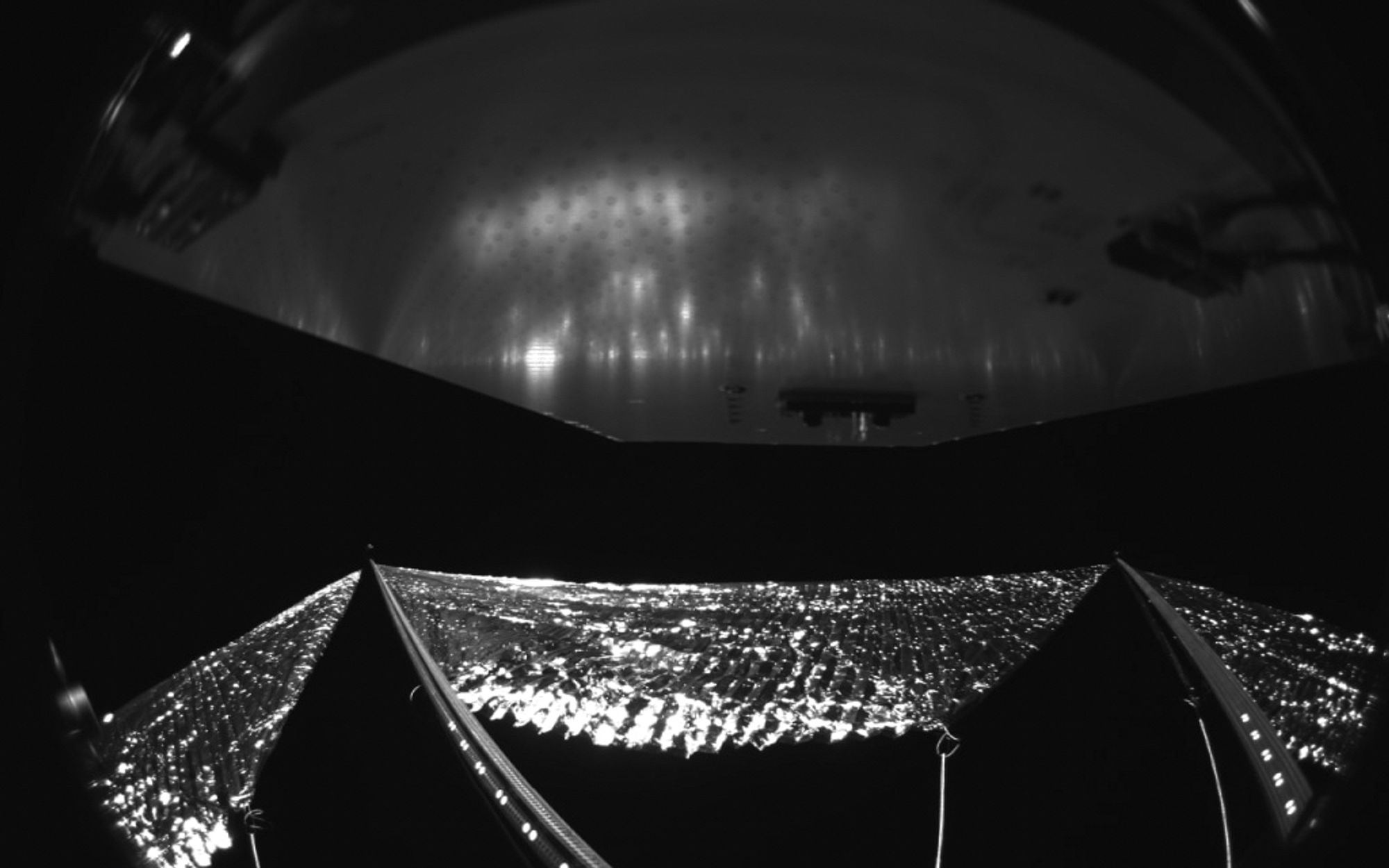 The Advanced Composite Solar Sail System has four black-and-white wide-angle cameras, centrally located aboard the spacecraft. Near the bottom of the photo, the view from one camera shows the reflective sail quadrants supported by composite booms. At the top of the photo is the back surface of one of the spacecraft’s solar panels. The five sets of markings on the booms close to the spacecraft are reference markers to indicate full extension of the sail. The booms are mounted at right angles, and the solar panel is rectangular, but appear distorted because of the wide-angle camera field of view. Credit: NASA