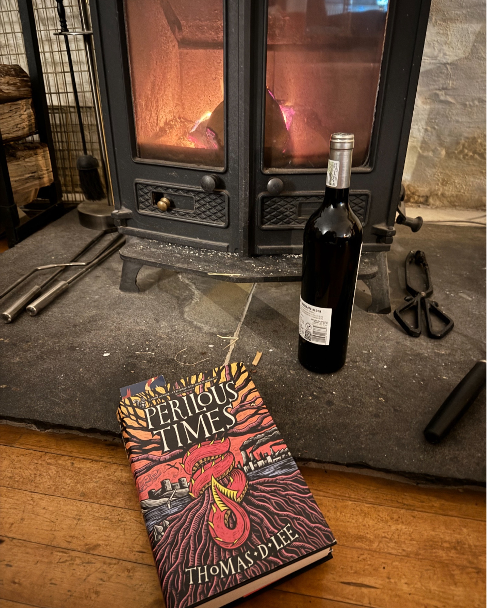 A somewhat untidy and dusty fireplace, with a lit stove and a bottle of red wine in front of it. Leaning against the fireplace is a copy of Perilous Times by Thomas D Lee. It is an orange and red cover with a red dragon tail wrapped around a tree.