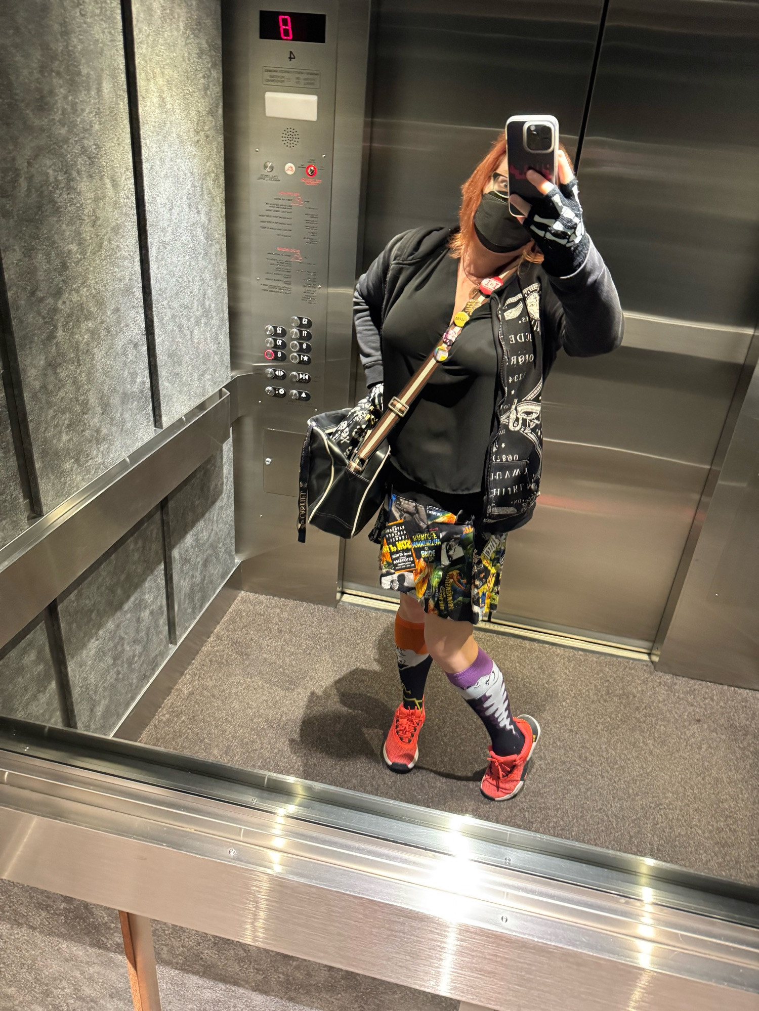 A self-portrait in a mirrored elevator. I am wearing a black, sheer top with my Ouija board hoodie over it, thrown open wide. My skirt is more fun. It is an amalgam of old school movie posters with Karloff and Lugosi heavily featured. There are ones for The Bride of Frankenstein and Son of Frankenstein amongst others. The colours are mainly black, orange, and green. I am wearing mismatched knee socks, but they are actually quite matched as the orange one features Frankenstein’s monster and the purple one features the Bride of Frankenstein. I am sporting my bright orange sneakers to match. The skelly gloves are on, too, this morning, dear alt text reader, as it is coooooold out there (don’t forget your booties!). 