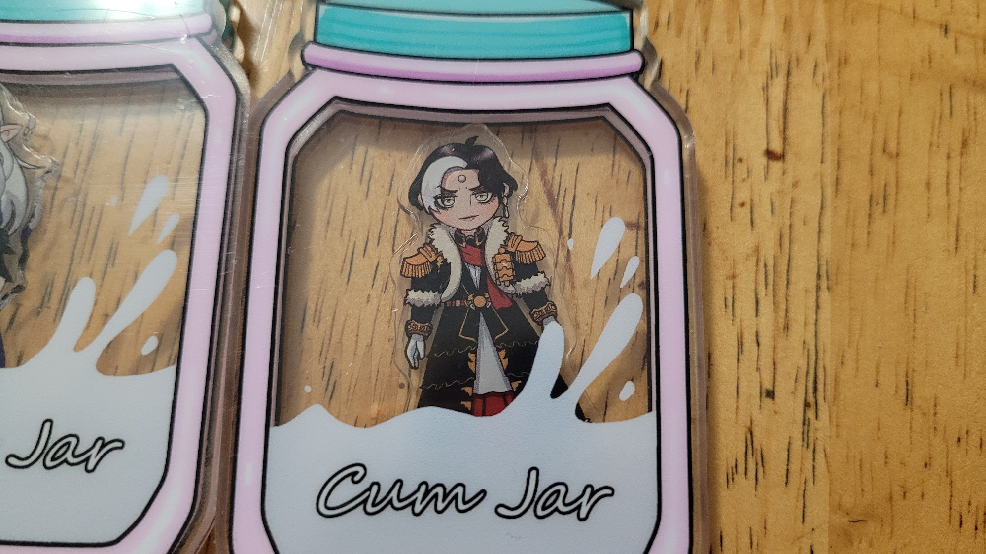 Description: an acrylic shaker keychain charm in the shape of a jar with a smaller charm of the character Emet-Selch from the video-game Final Fantasy 14 inside of it. The jar has the words "cum jar" on it with a white splash inside. 