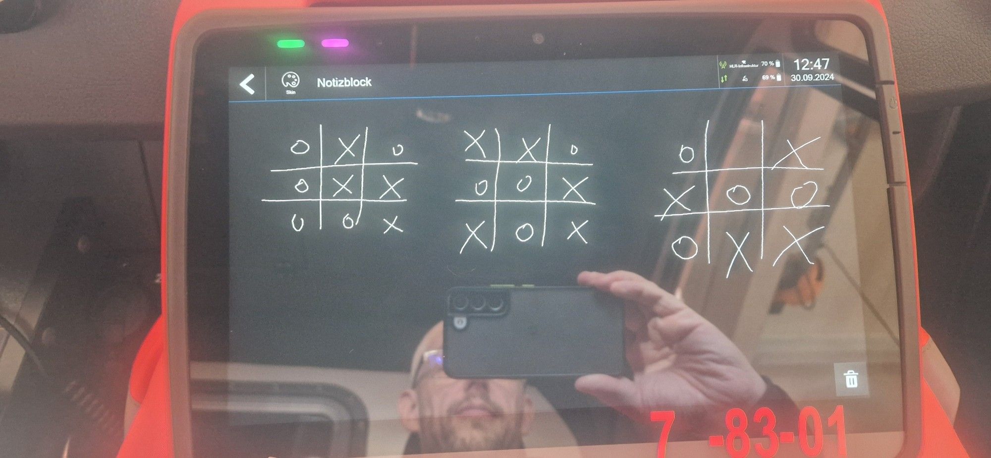 Das Bild zeigt erneut ein Tablet, auf dem die Notizblock-App geöffnet ist. Auf dem Bildschirm sind drei Runden eines Tic-Tac-Toe-Spiels zu sehen, jeweils mit Kreuzen („X“) und Kreisen („O“). Jedes Spielfeld zeigt ein unentschiedenes Spiel, da keines der beiden Symbole eine vollständige Reihe gebildet hat. Auf dem Bildschirm spiegelt sich eine Person, die ein Smartphone hält, welches die Reflexion der Szene aufnimmt. Das Tablet befindet sich wie zuvor auf den Knien der Person, die eine rote Hose trägt. Im Hintergrund sind Teile des Fahrzeuginnenraums erkennbar.