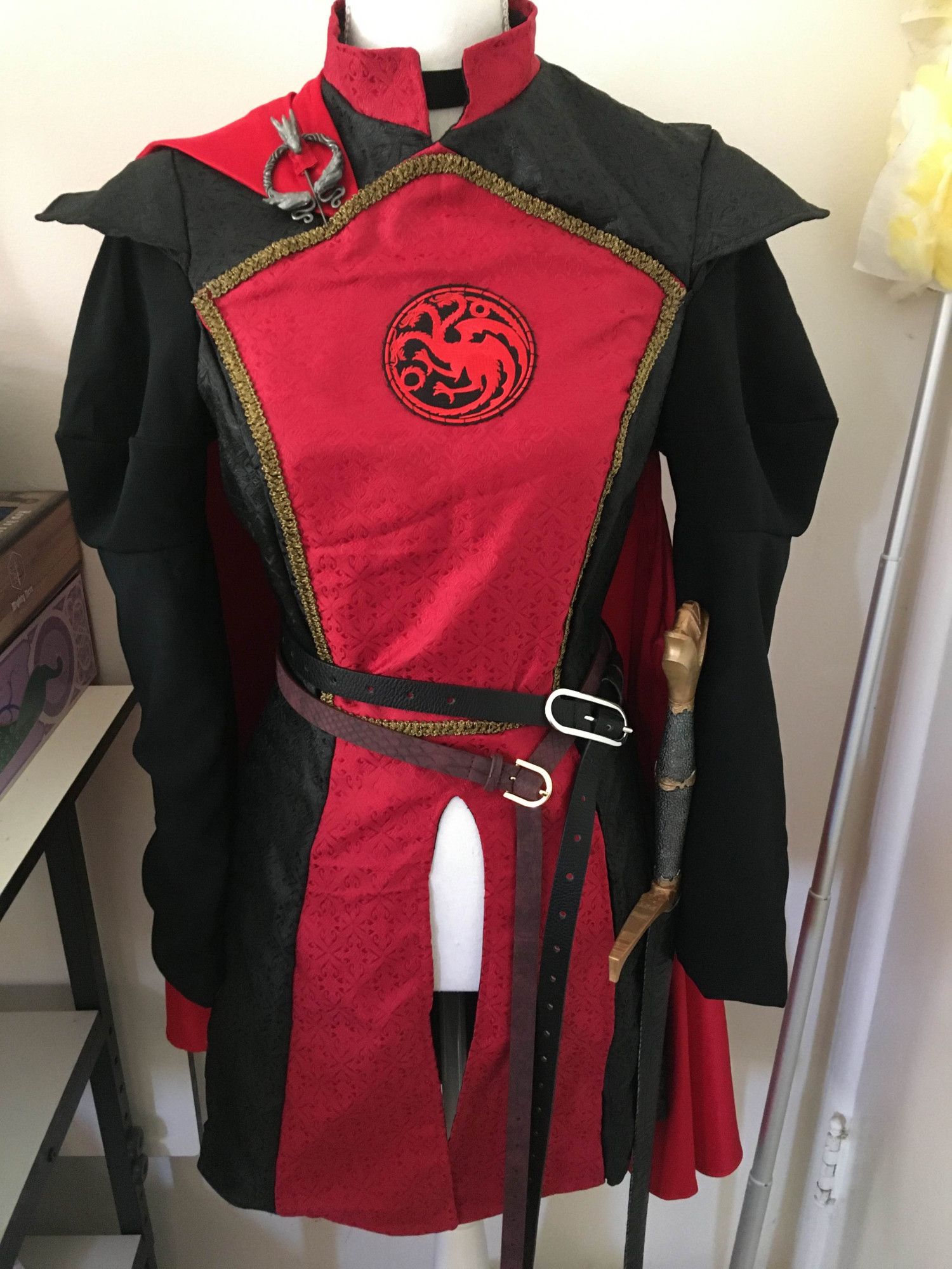 A black and red brocade surcoat with lantern sleeves, a red shoulder cape, and a sigil of House Targaryen sewed to the center panel. There are two belts wrapped around the waist, with a 3D printed sword attached