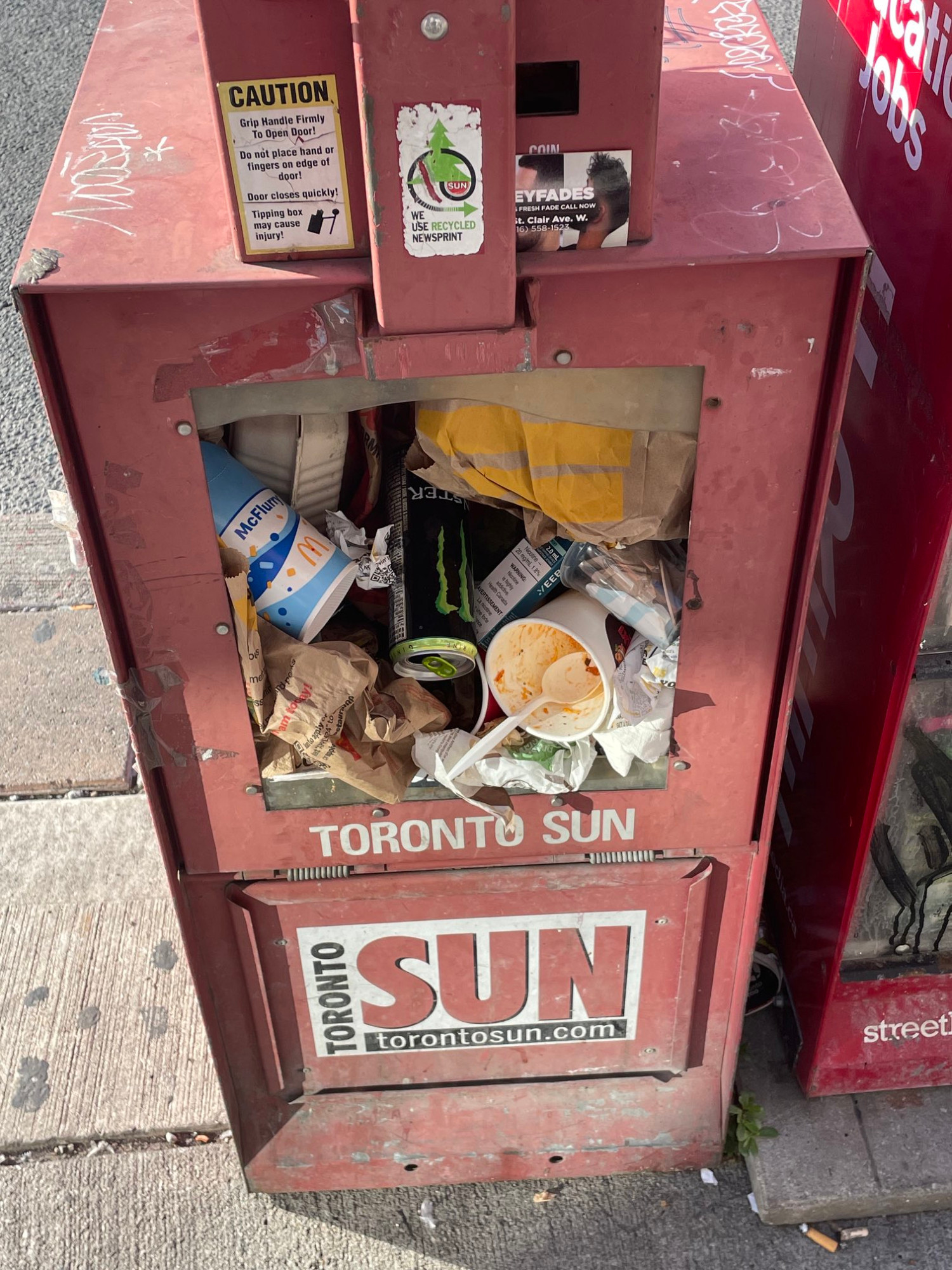 An old sidewalk newspaper dispenser, now filled with trash instead of papers