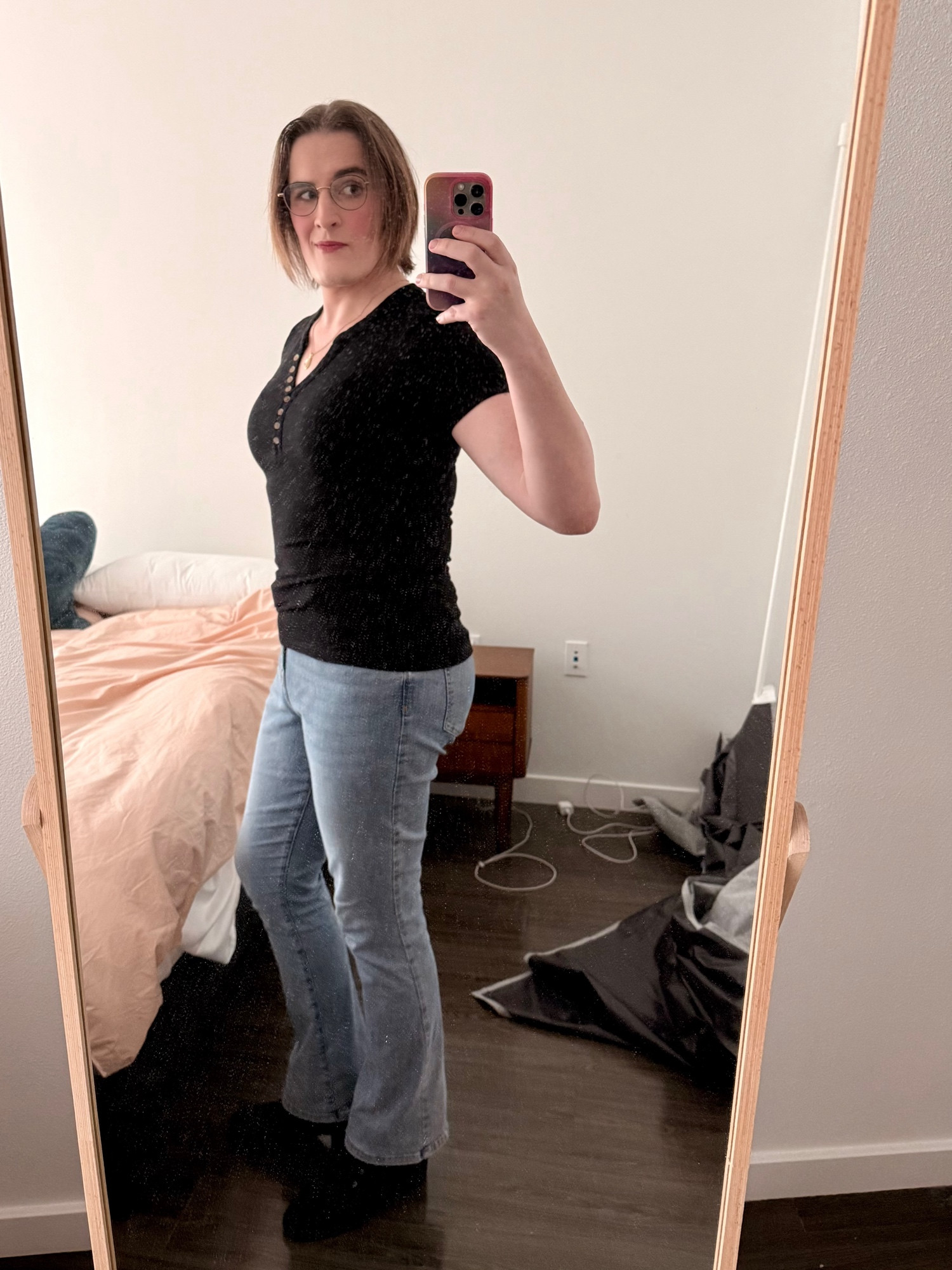A white woman with brown hair stands before a bedroom floor-length mirror. She’s wearing a black shirt, light-wash boot-cut jeans, and black boots with a little heel.
