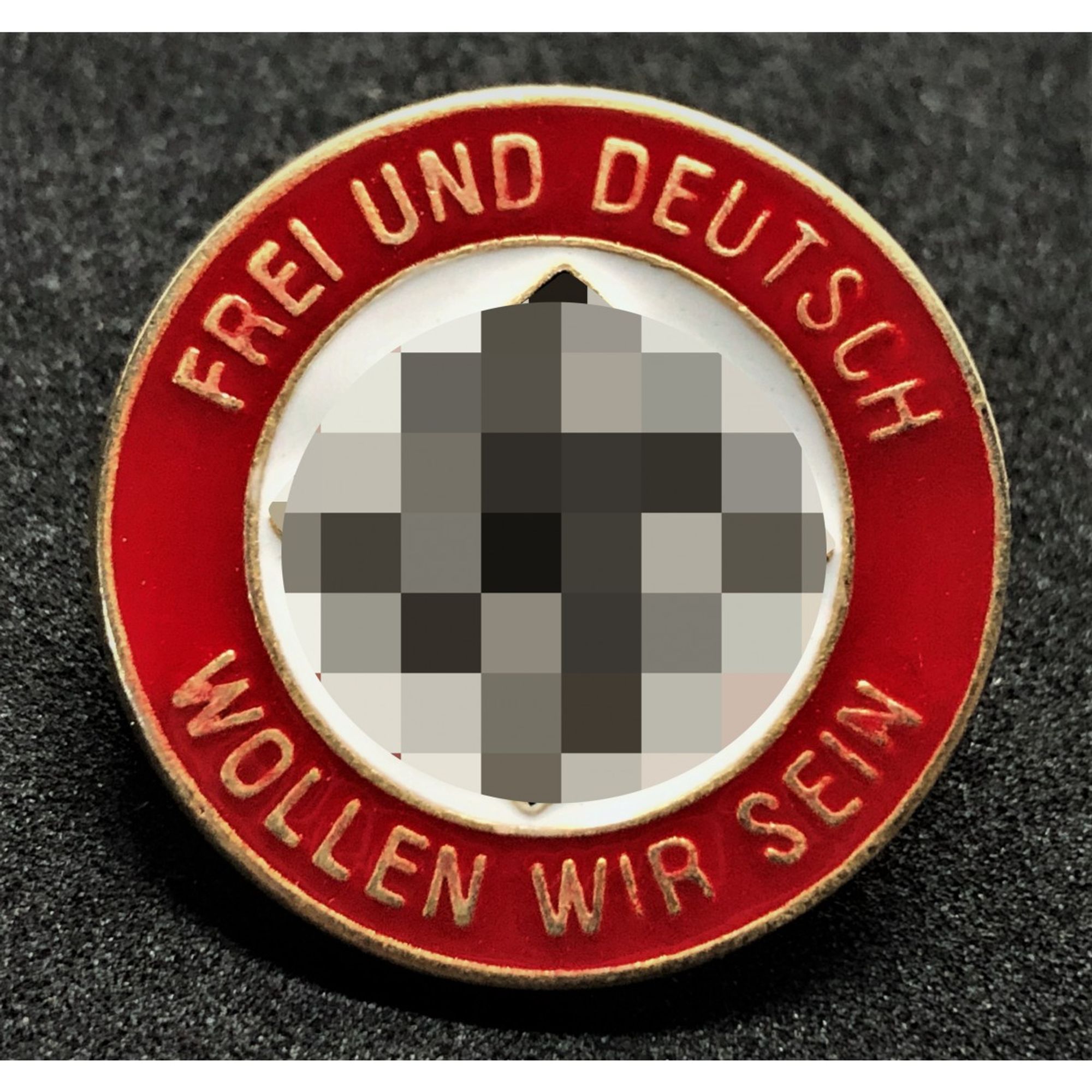 Metallabzeichen der Nazis mit der Aufschrift "FREI UND DEUTSCH WOLLEN WIR SEIN" am Rand und in der Mitte ein verpixeltes Hakenkreuz.