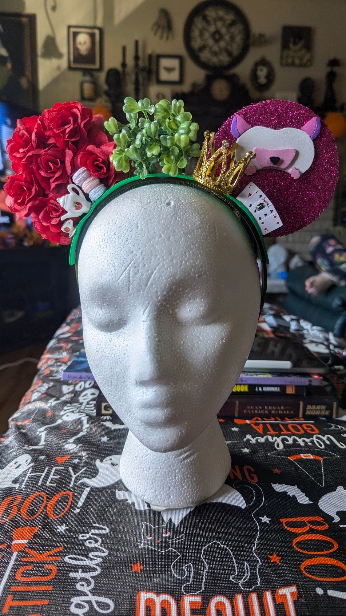 A pair of handcrafted Mickey ears sit on a white mannequin head. One ear is made of red silk roses, the other is a pink rolled-up hedgehog made out of layers of craft foam. Trinkets including miniature tea wear, a crown, and playing cards adorn the band