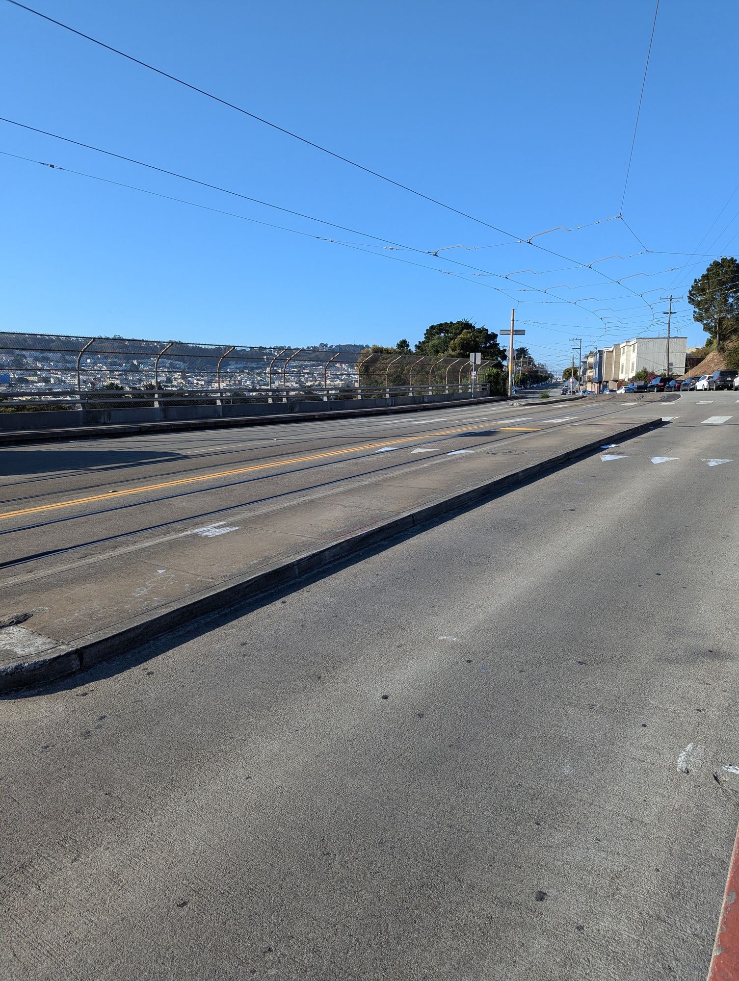 A low floor light rail boarding platform next to a car lane on one side and railroad tracks on the other. Behind it are a second similar platform for the other direction of travel, and a few houses.