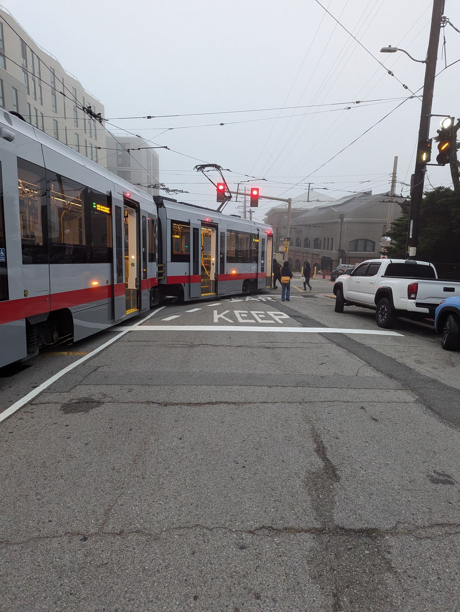 A light rail vehicle stopped on a city street. It is blocking the traffic lane and is about to turn into a rail yard. This is in foggy morning light.