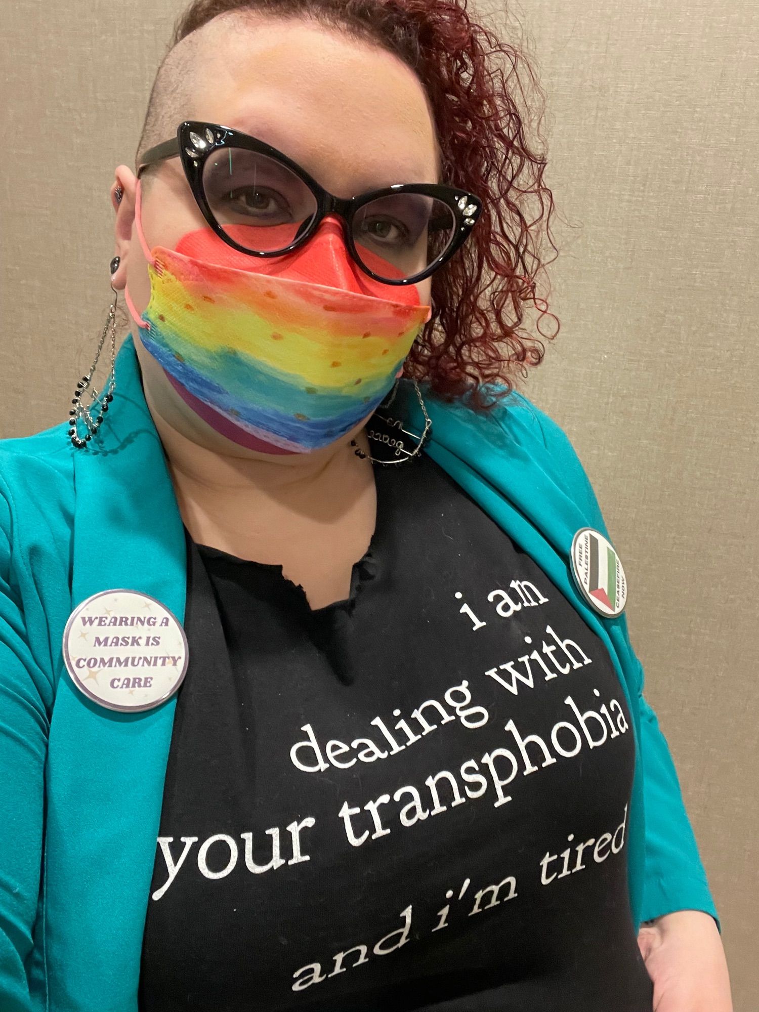 Red head white nonbinary femme in a rainbow mask. A shirt says I am dealing with your transphobia and I am tired. One button says masking is community care and the other is in support of Palestine