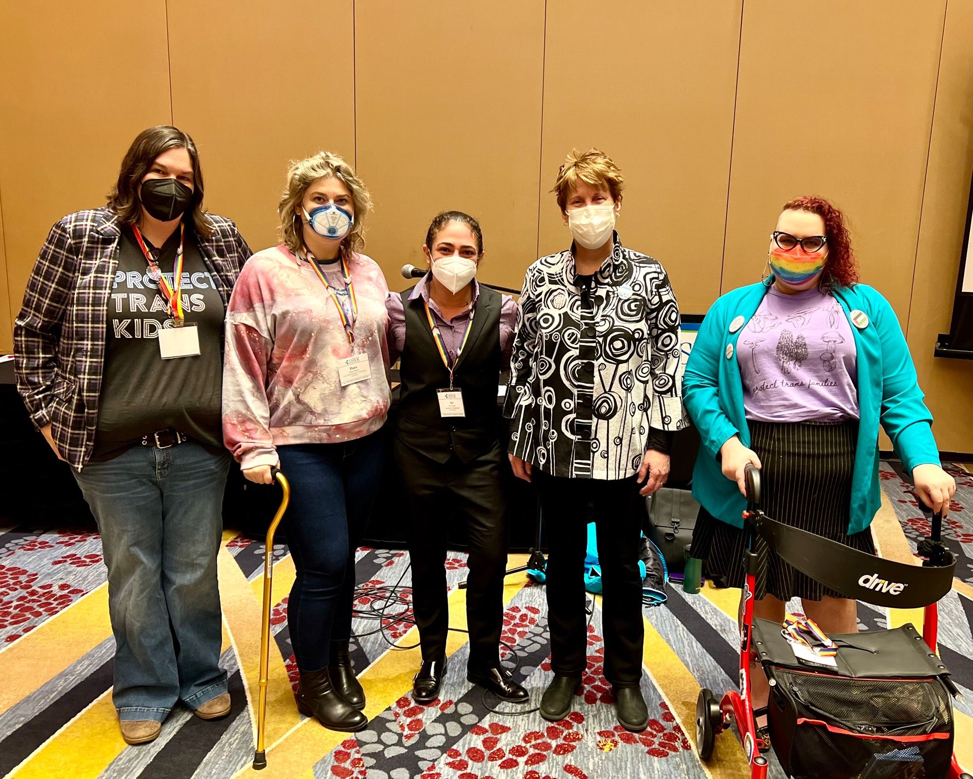 5 queer folks standing in a hotel conference presentation room, masking