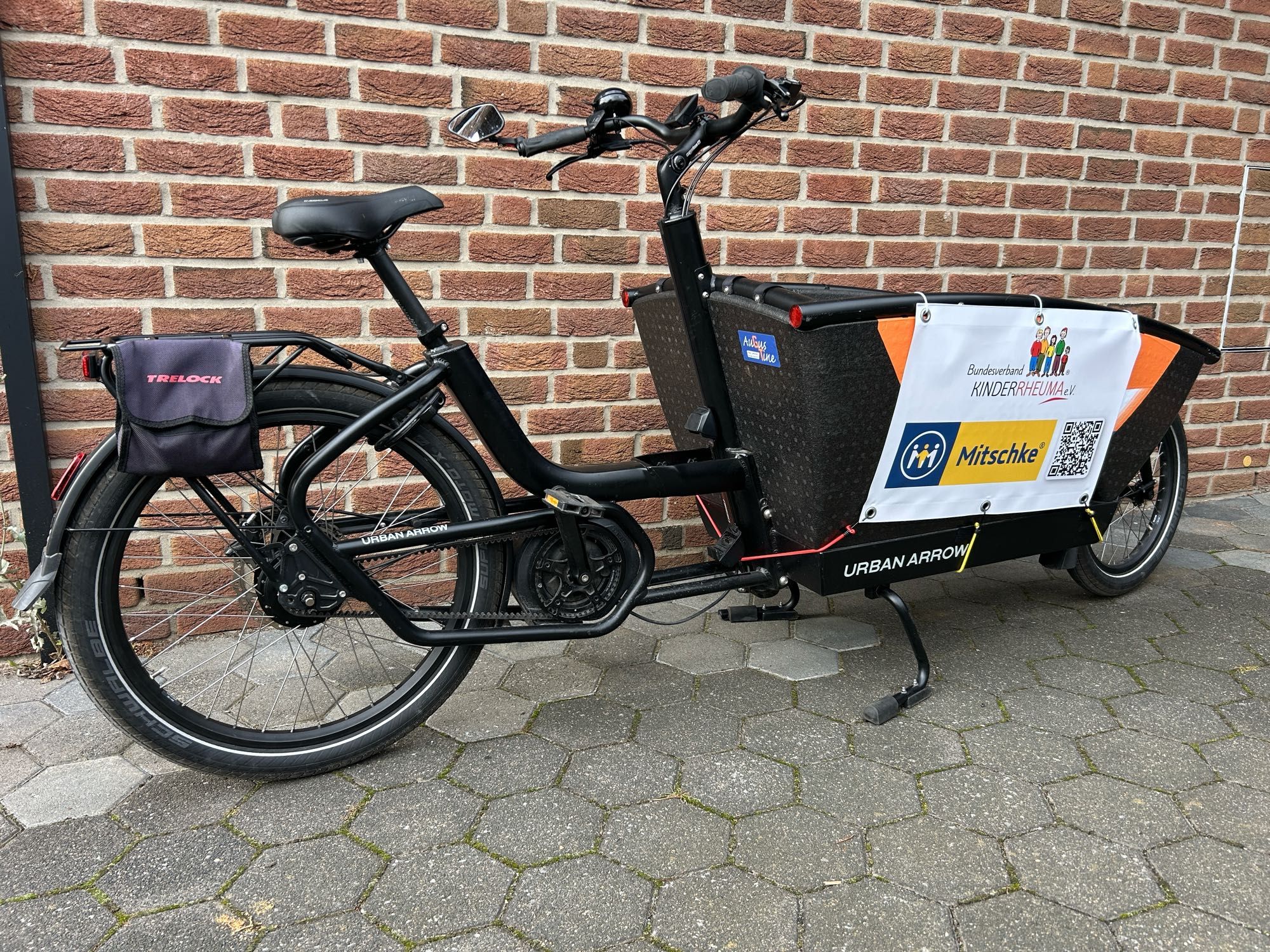 Das Bild zeigt ein Lastenfahrrad der Marke Urban Arrow, das an einer Ziegelsteinmauer lehnt. Das Fahrrad hat einen großen schwarzen Transportbehälter vorne. Auf dem Behälter ist ein Banner befestigt, auf dem "Bundesverband KINDERRHEUMA e.V." und "Mitschke" steht, zusammen mit einem QR-Code. Am hinteren Teil des Fahrrads ist eine kleine Tasche mit der Aufschrift "TRELOCK" angebracht. Das Fahrrad steht auf einem gepflasterten Boden.