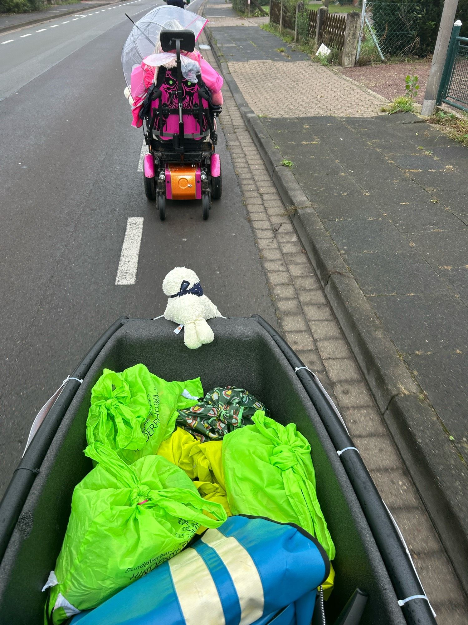 Das Bild zeigt eine Straße mit einem Bürgersteig auf der rechten Seite. Auf der Straße fährt ein elektrischer Rollstuhl, der mit einem durchsichtigen Regenschirm und einer pinkfarbenen Abdeckung ausgestattet ist. Auf der Rückseite des Rollstuhls ist ein orangefarbenes Element zu sehen. Vor dem Rollstuhl befindet sich ein Anhänger oder eine Art Transportbehälter, der mit mehreren leuchtend grünen Taschen und einer blauen Tasche gefüllt ist. Auf dem Behälter sitzt ein kleines weißes Stofftier. Die Umgebung wirkt wie eine Wohngegend mit einem Zaun und einigen Pflanzen am Straßenrand.