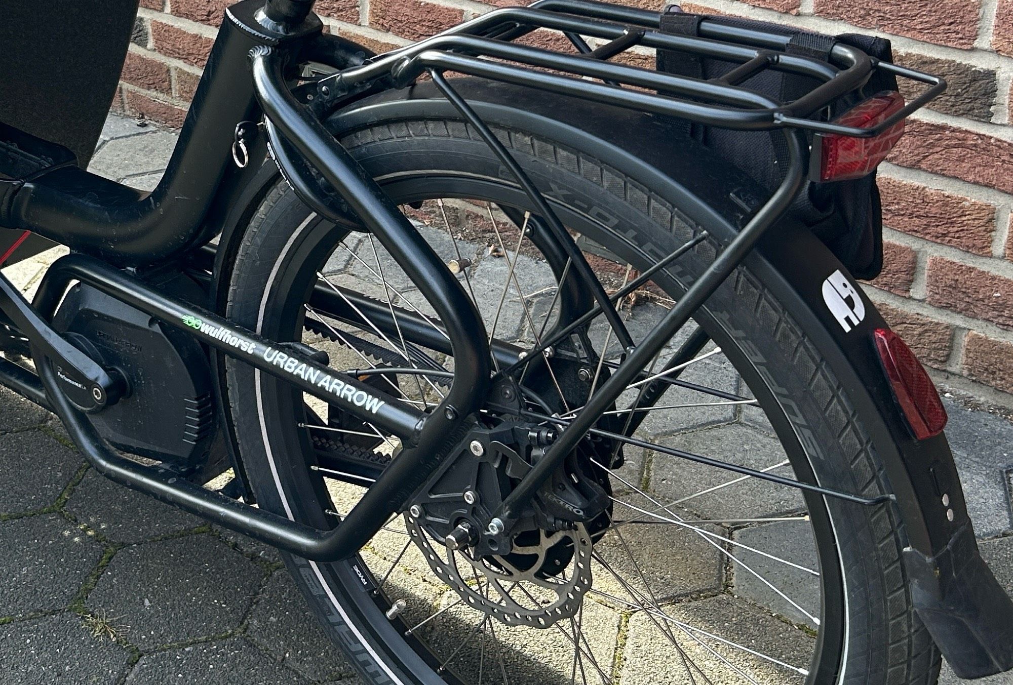 Das Bild zeigt das hintere Teil eines schwarzen Fahrrads, das an einer Ziegelsteinmauer lehnt. Es handelt sich um ein Lastenfahrrad der Marke "Urban Arrow". Man sieht den Gepäckträger über dem Hinterrad, das mit einem Schutzblech ausgestattet ist. Am Schutzblech ist ein Rücklicht angebracht und ein Aufkleber der einen weinenden Elefanten zeigt. Der Rahmen des Fahrrads ist robust und für den Transport von Lasten ausgelegt. Der Boden besteht aus Pflastersteinen.