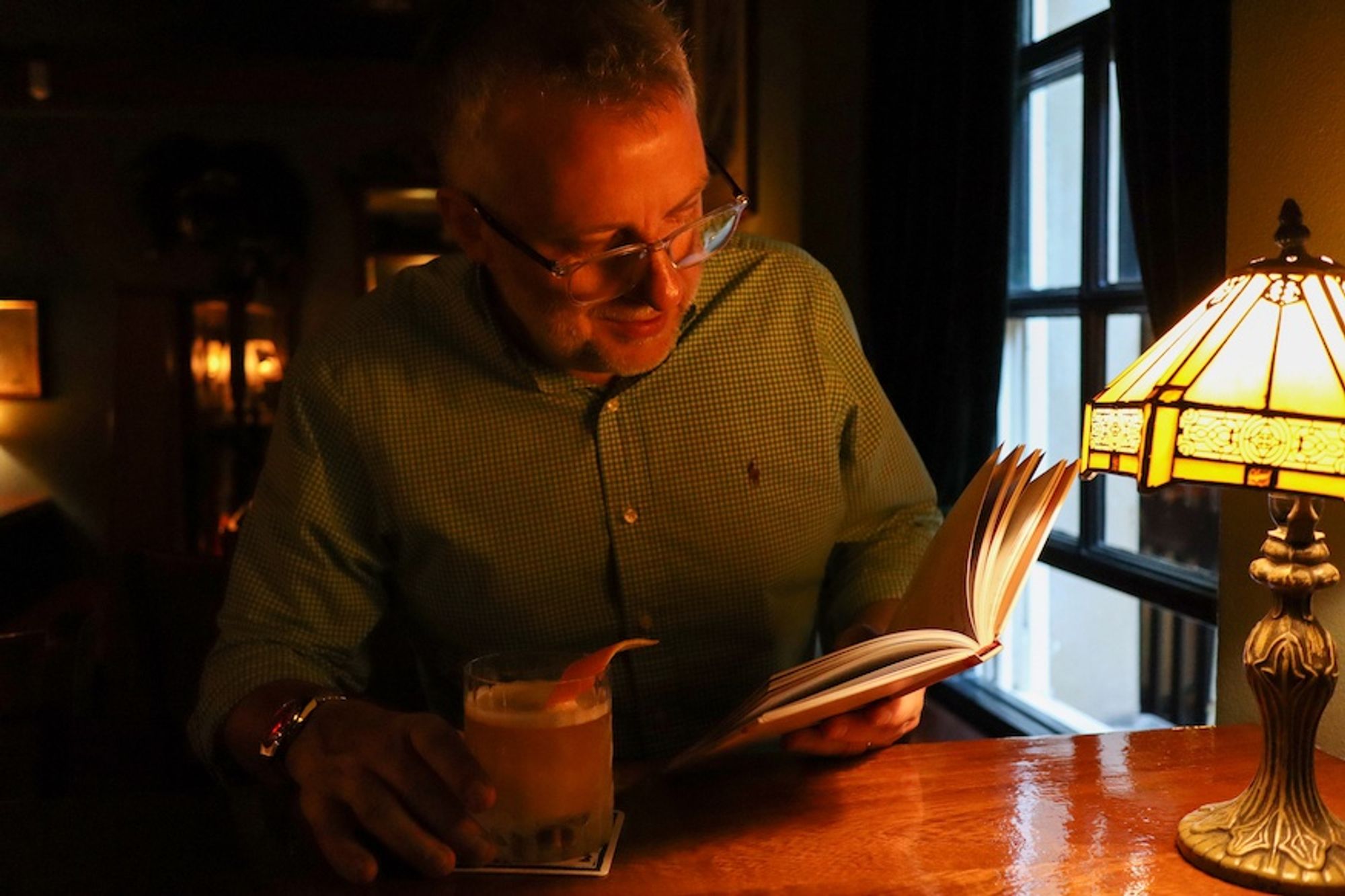 A man sits in a dimly lit speakeasy with a sidecar cocktail and a book. Photo by Rhian Drinkwater.