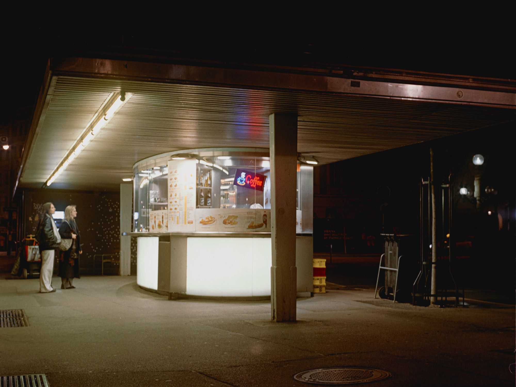 a coffee stand in vienna, shot at about 4:00AM on a friday night, it shows two women looking at the brightly illuminated coffee stand