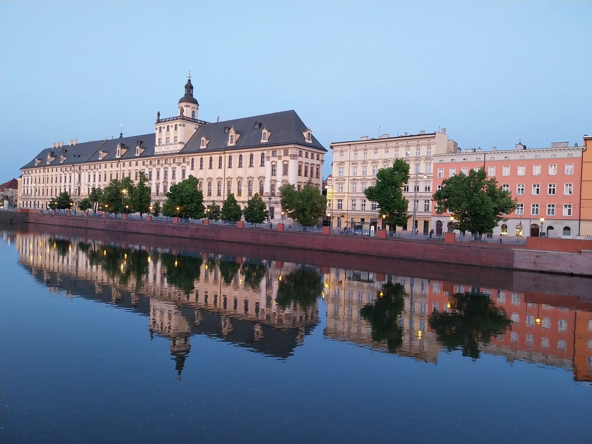 Im abendlichen Breslau spiegelt sich die Häuserfassade der Universität Breslau, ein langgezogenes 3-stöckiges Gebäude mit weißer Fassade, im Fluss Odra (Oder)