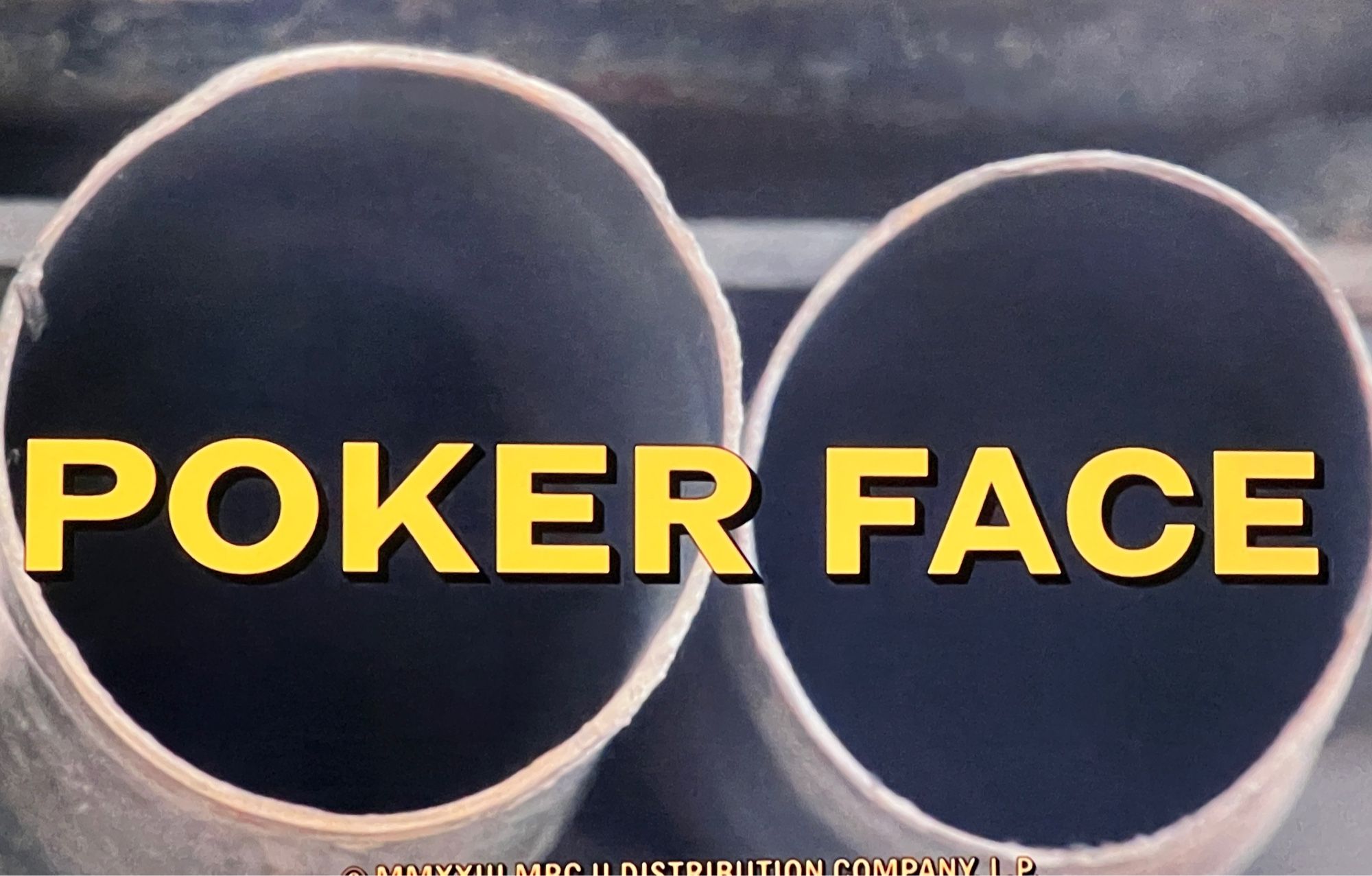 Yellow lettering spelling out the words Poker Face in similar font to previous image. In background two large cylindrical pipes.