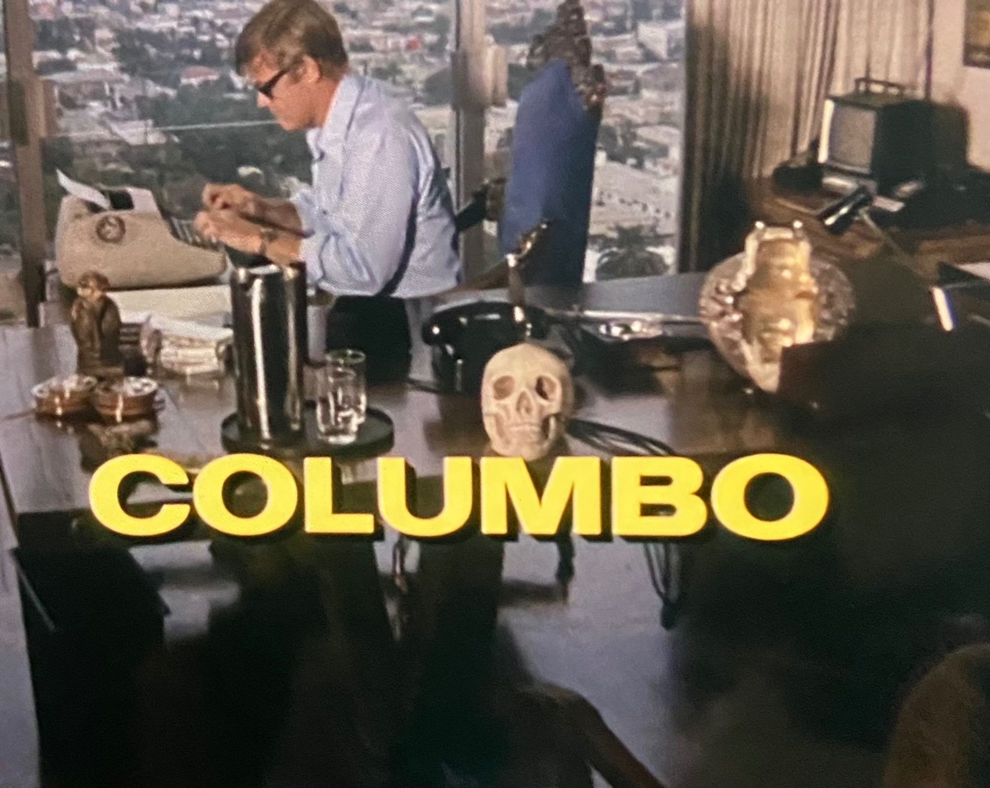 Yellow letters spell out Columbo in front of a scene of a white make typing on an old typewriter by a desk covered with random artifacts including a skull and a rotary phone.