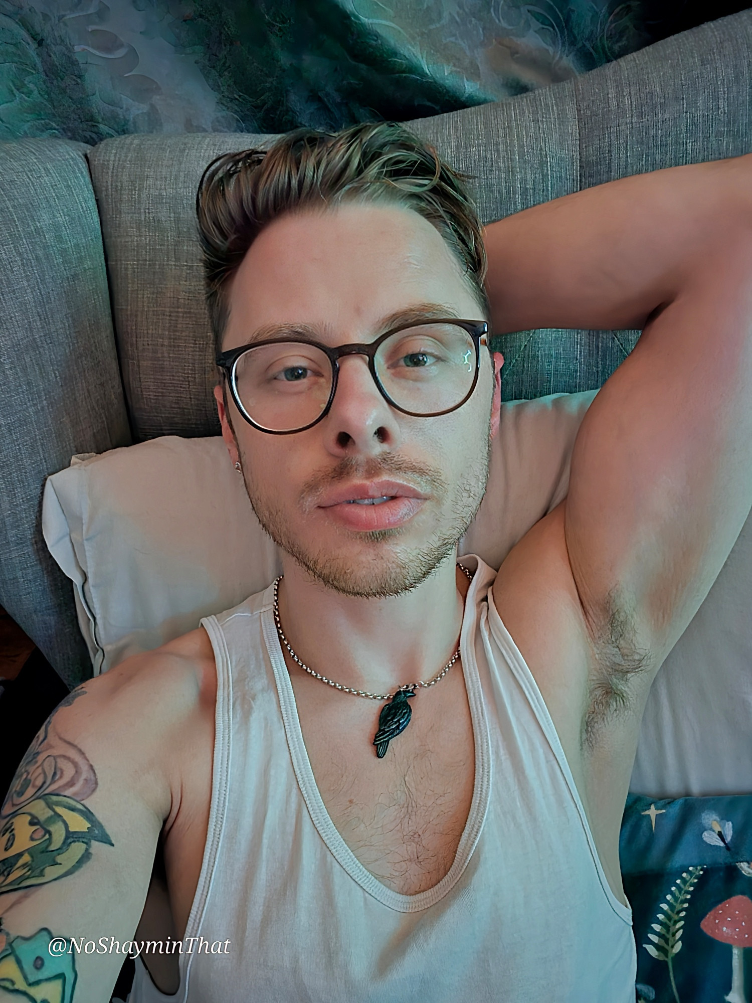 It's me, lying on a comfy pillow against a grey headboard. I'm wearing my glasses and a white tank top, and you can see the chain with my favorite crow pendant. I've got my arm tucked behind my head, and you can see a bit of my tattoos on my left arm.
