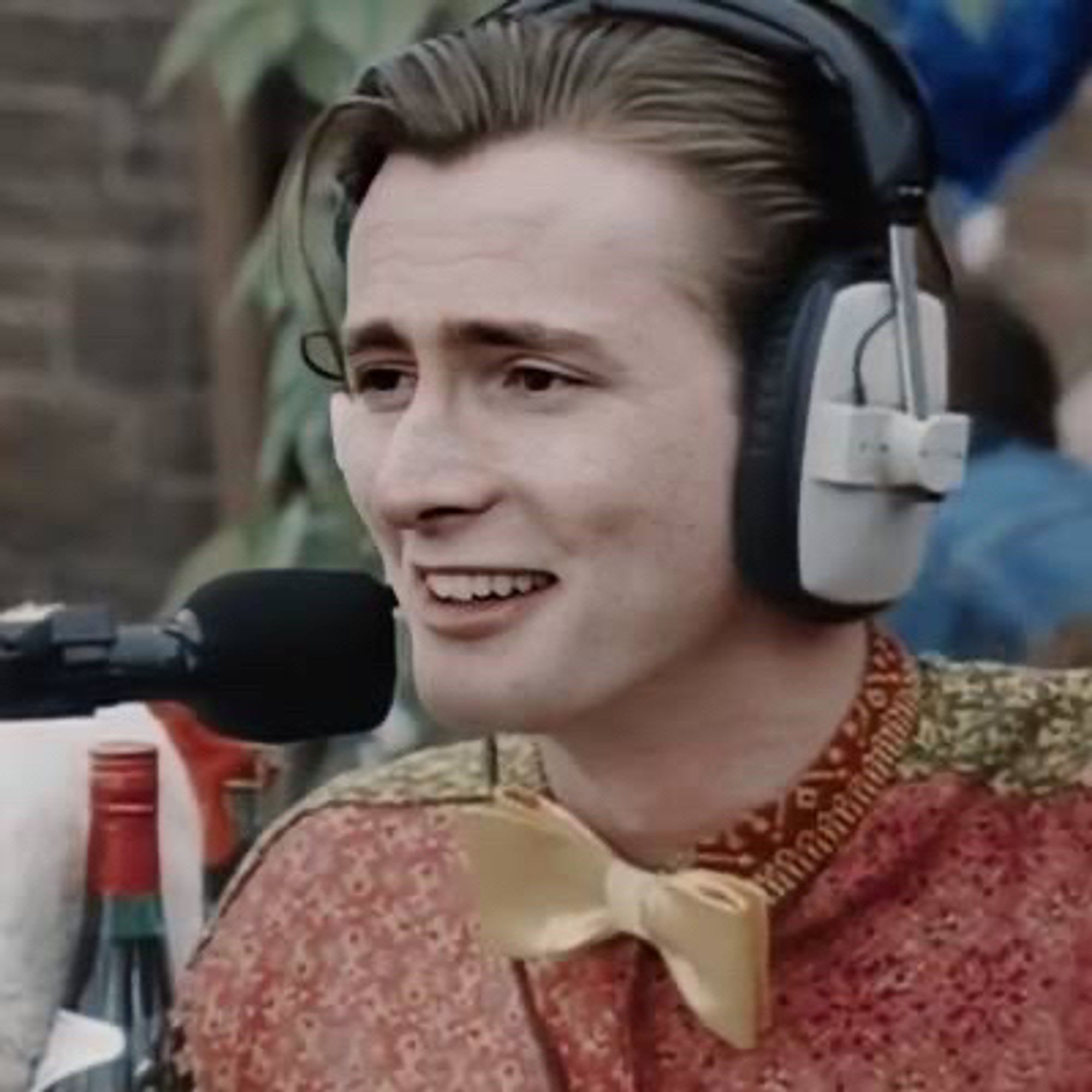 Young David Tennant as Campbell Bain. His hair is straight, brown and has blond lights. He is wearing a gold bow tie and a red and green patterned shirt. His wearing a headphone and speaking on a microphone.