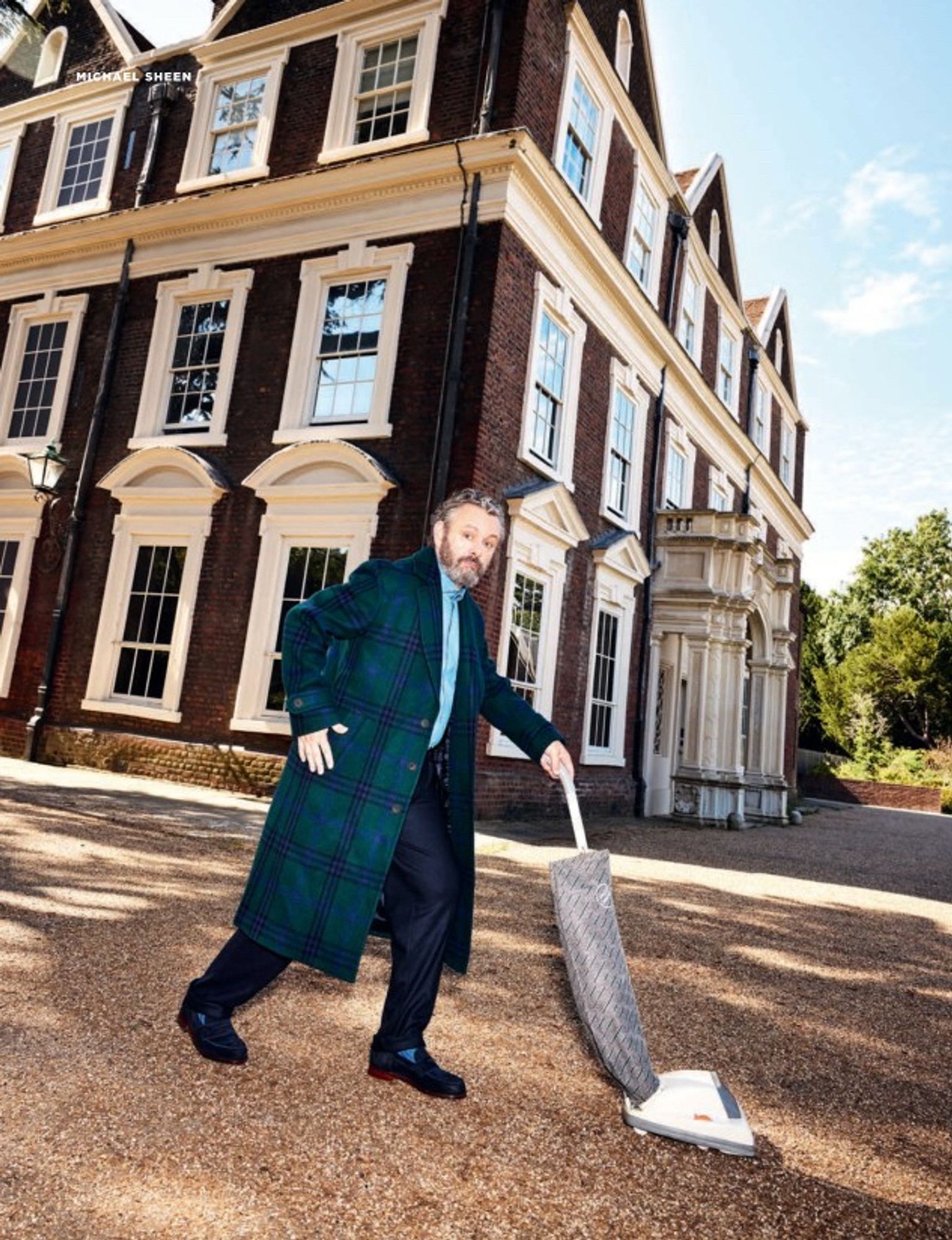 Michael Sheen using a vacuum cleaner outside a big house