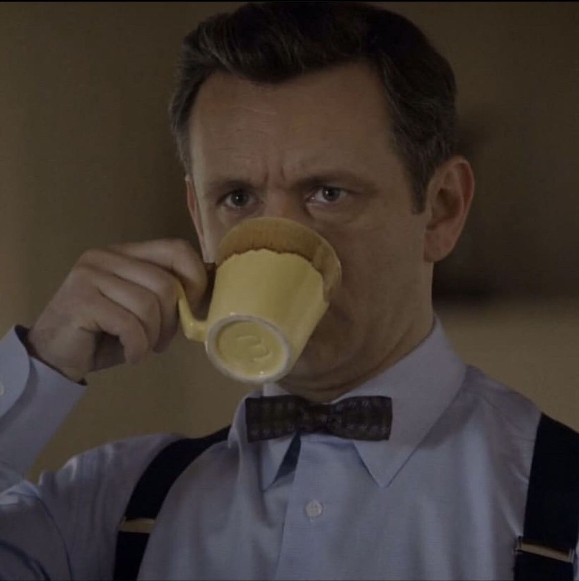 Michael Sheen as Bill Masters from the tv show Masters of Sex. He’s wearing a light blue social shirt with a dark red bow tie and suspenders. He is drinking from a yellow mug that is covering the bottom half of his face. His eyebrows are drawn together and he is looking at something in front of him