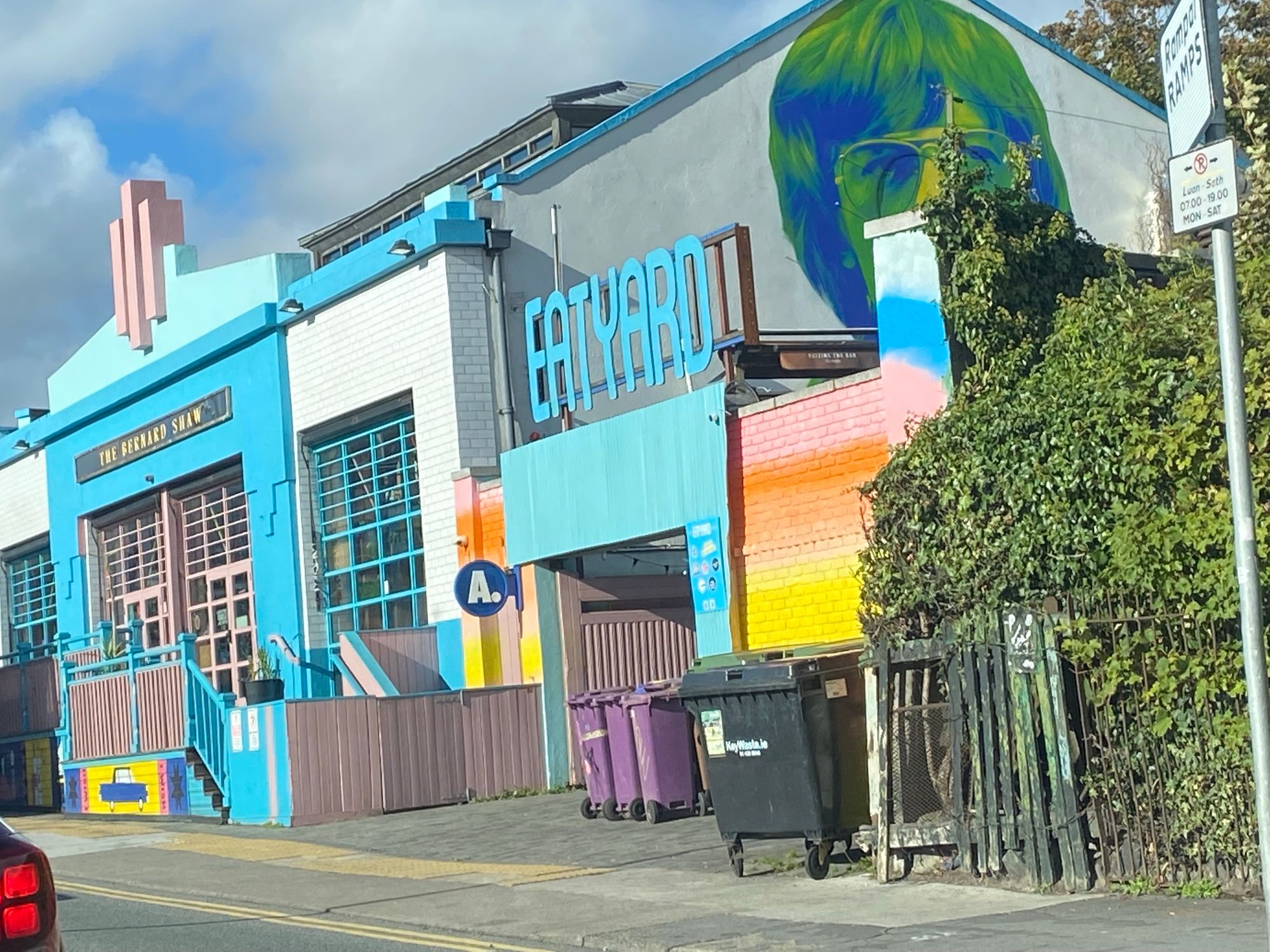 Ein farbenfrohes Gebäude mit dem Schild "EAT YARD" neben einem Wandbild einer Person mit Brille. Die Fassade hat leuchtende Farben, und vorne befinden sich lila Mülleimer. Üppiges Grün verdeckt teilweise einen Zaun.


A  building featuring the sign "EAT YARD" alongside a mural of a person with glasses. The facade has vibrant colors, and there are purple trash bins in front. Lush greenery partially obscures a fence.