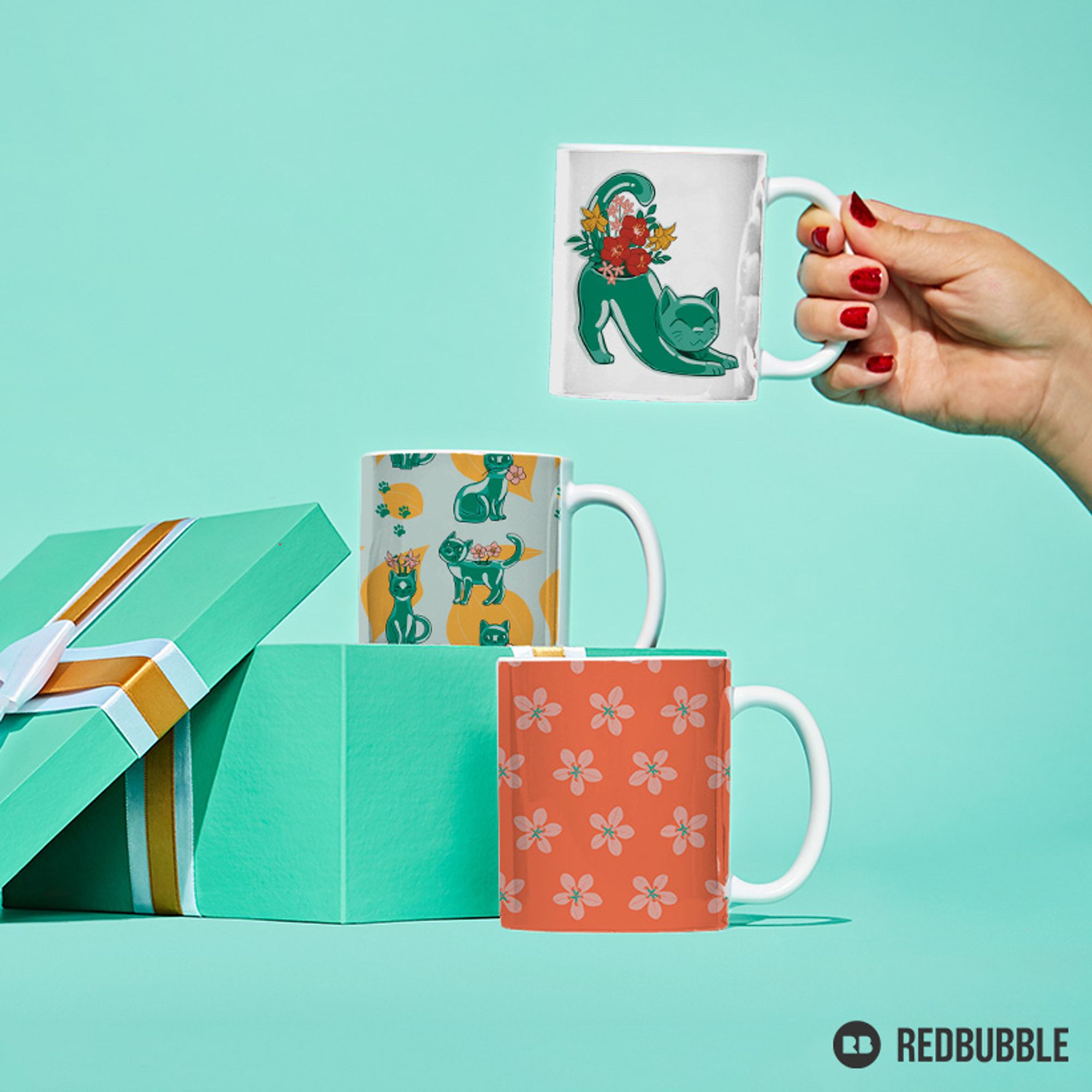 Three mugs with various patterns on them. A femme hand holds up one with a jade cat vase on it. One is in an open green present box. It has a pattern of jade cats on it. The third mug is in front of the box with a pattern of spring beauty flowers.