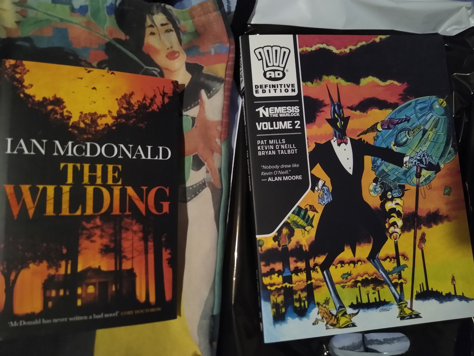 New novel by Ian McDonald - The Wilding - a big house, beneath trees, with lots of autumnal/fire colours round the dark parts. And the 2nd in the Definitive series of Nemesis the Warlock collections from 2000AD - cover shows dapper, devilish alien, dressed in a suit, with a cane, and zeppelin hovering above industrial plant in background, colour tones similar to novel, orange and fiery.