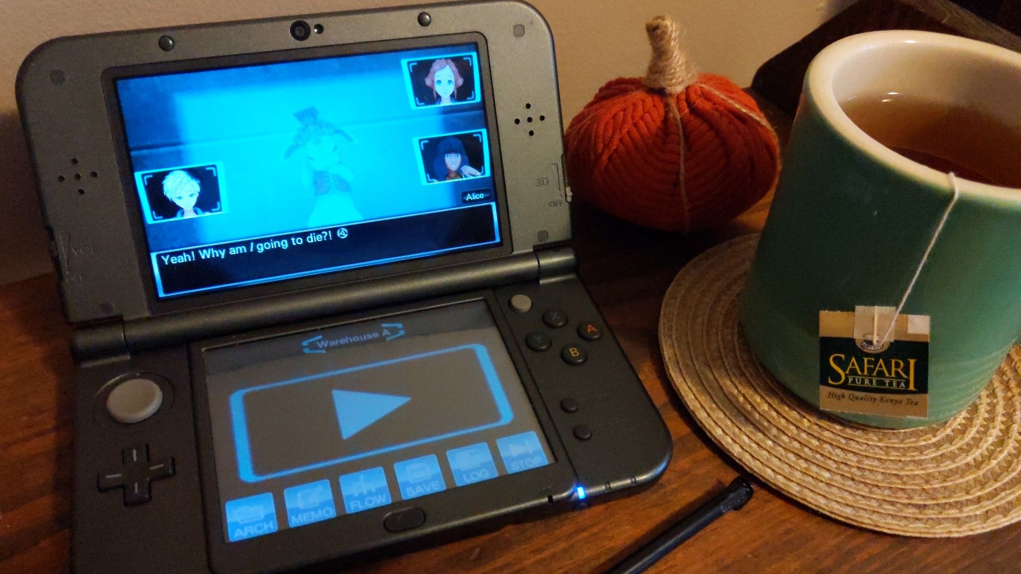 A 3DS video game system is on a side table along with a green bug of tea, a stylus, and a decorative pumpkin. On the screen is the game Virtue's Last Reward, with the speaking character exclaiming "Yeah! Why am *I* going to die?"