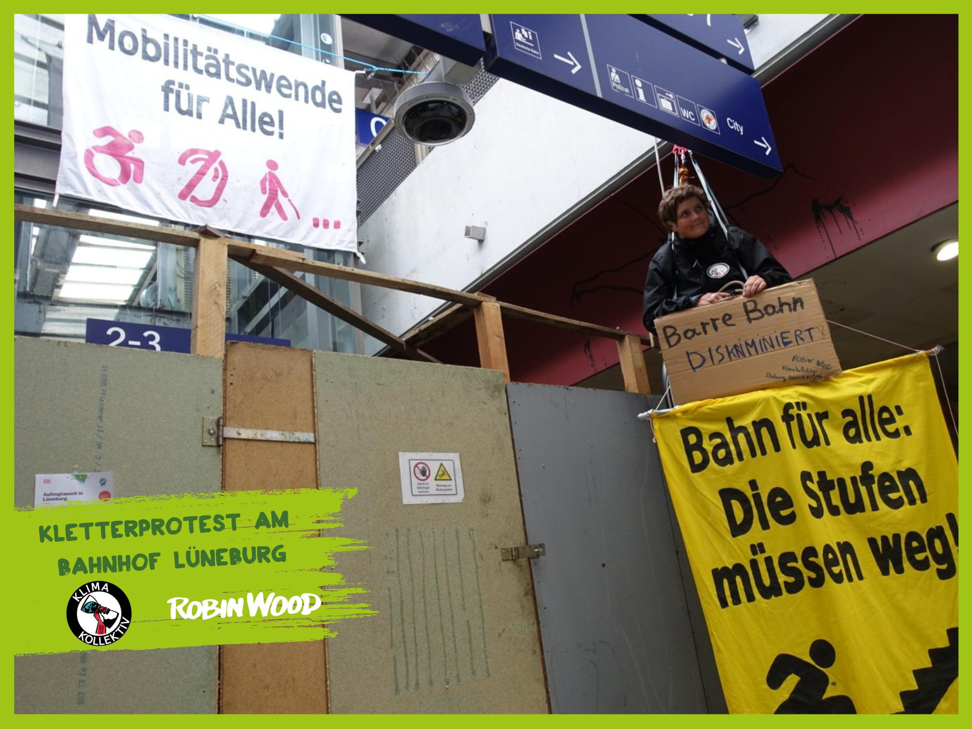 Eine ROBIN WOOD-Aktivistin seilte sich in ihrem Rollstuhl an der Bahnhofsunterführung ab. Sie hält ein Schild und Banner mit der Aufschrift "Barriere Bahn diskriminiert! Bahn für alle: Die Stufen müssen weg!" Andere Aktivist*innen hängten im Hintergrund ein Banner mit der Forderung "Mobilitätswende für Alle!" auf.